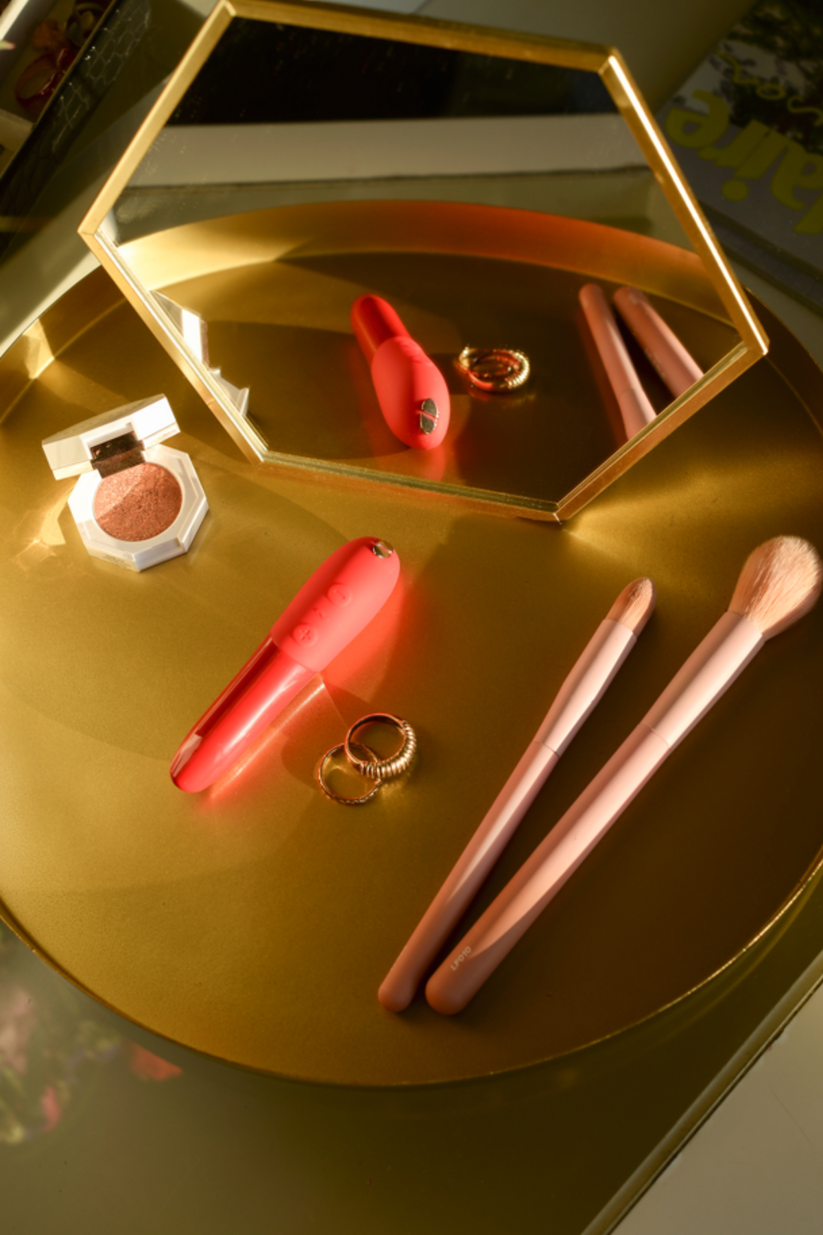 We-Vibe Tango X bullet vibrator in Cherry Red on a gold vanity tray beside makeup brushes, a compact and rings, with a mirror