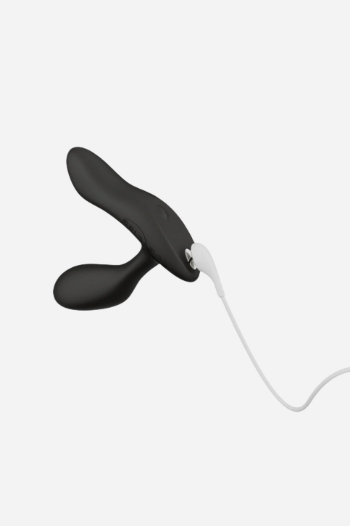 Black We-Vibe Vector+ with its white magnetic USB charging cable attached to the base, showing the rechargeable design on light grey