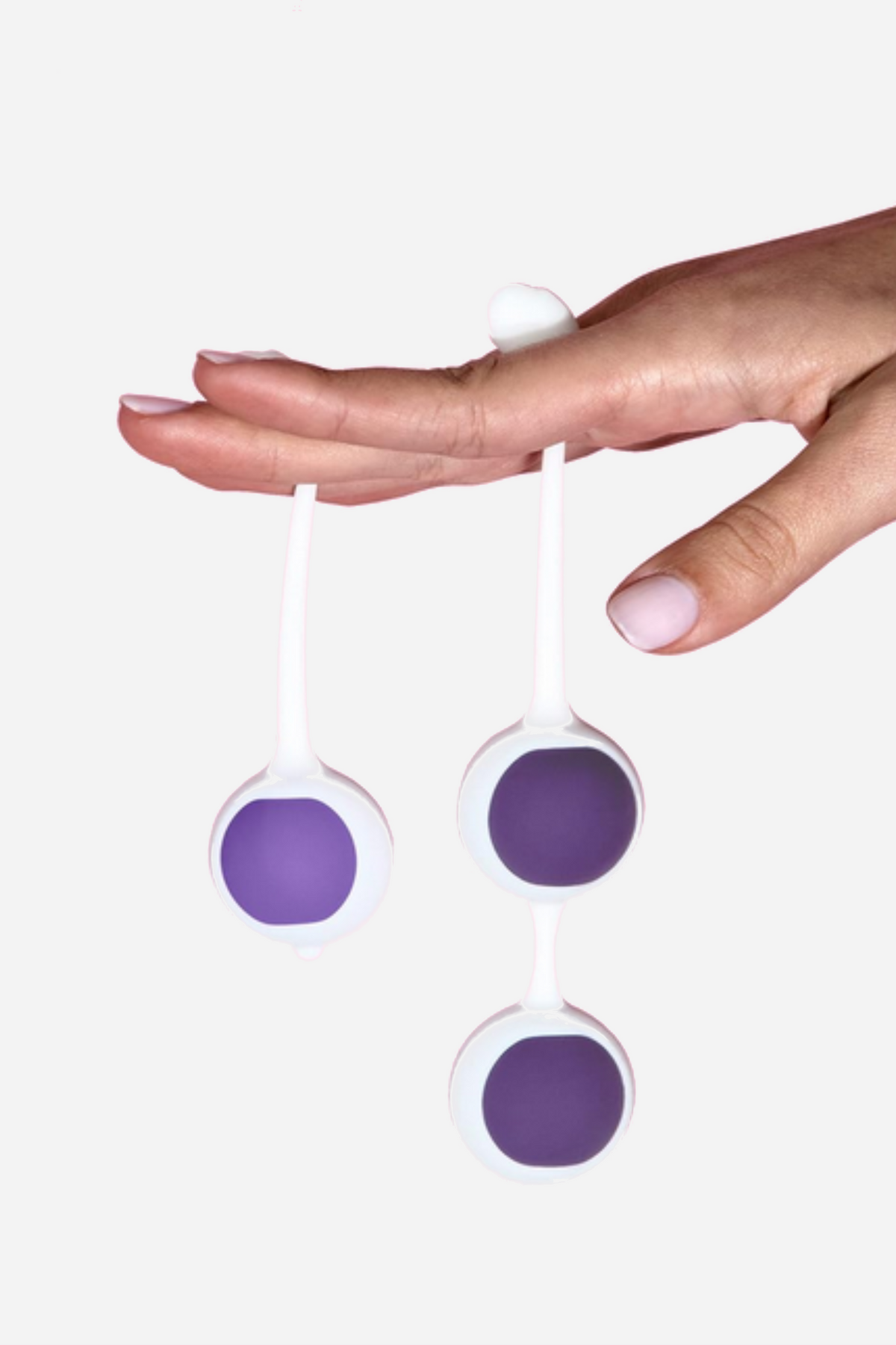 A hand with fingers extended holds three purple and white kegel balls dangling from their retrieval cords, against a soft pink background.
