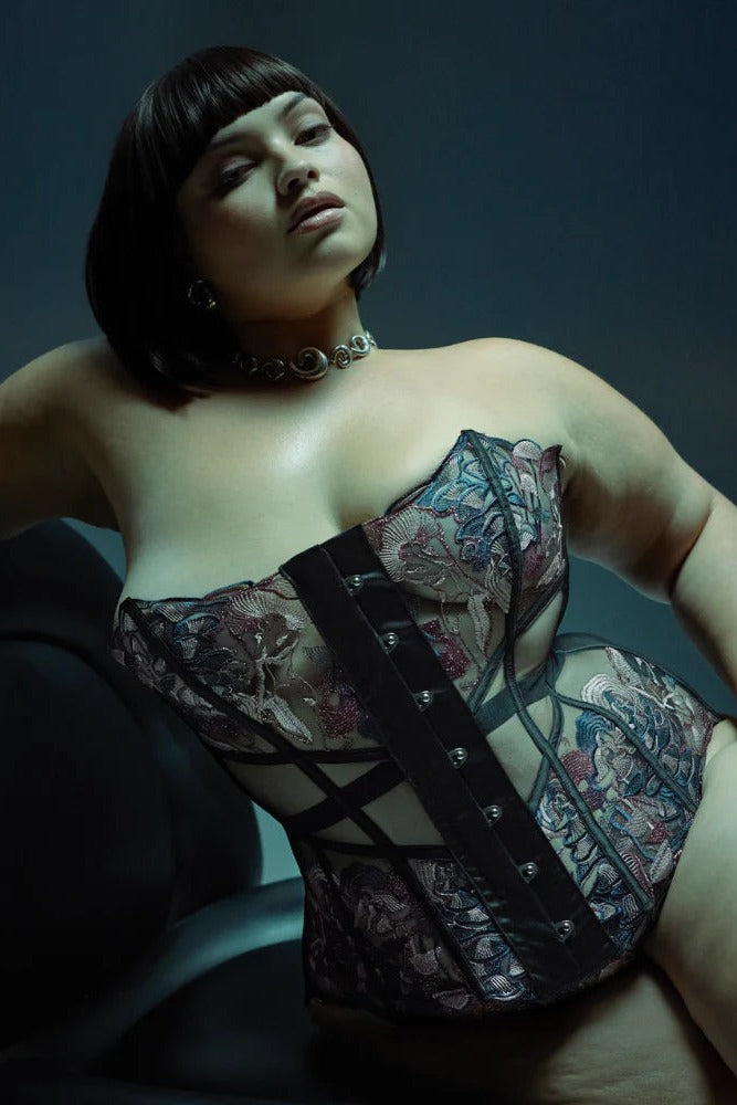Curvy model with a black bob reclining in a chair in the Wild Chanterelle Dreams corset, showing the embroidered cups and boning against a teal backdrop