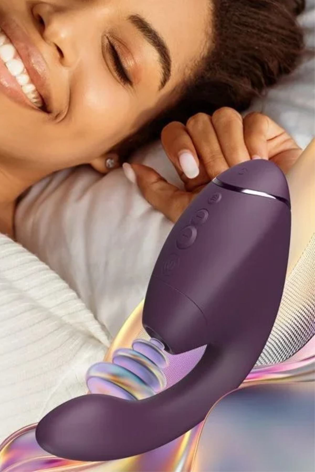 Smiling woman lying in bed holding the dark purple Womanizer Next Duo rabbit vibe, with its Pleasure Air head and G-spot arm visible
