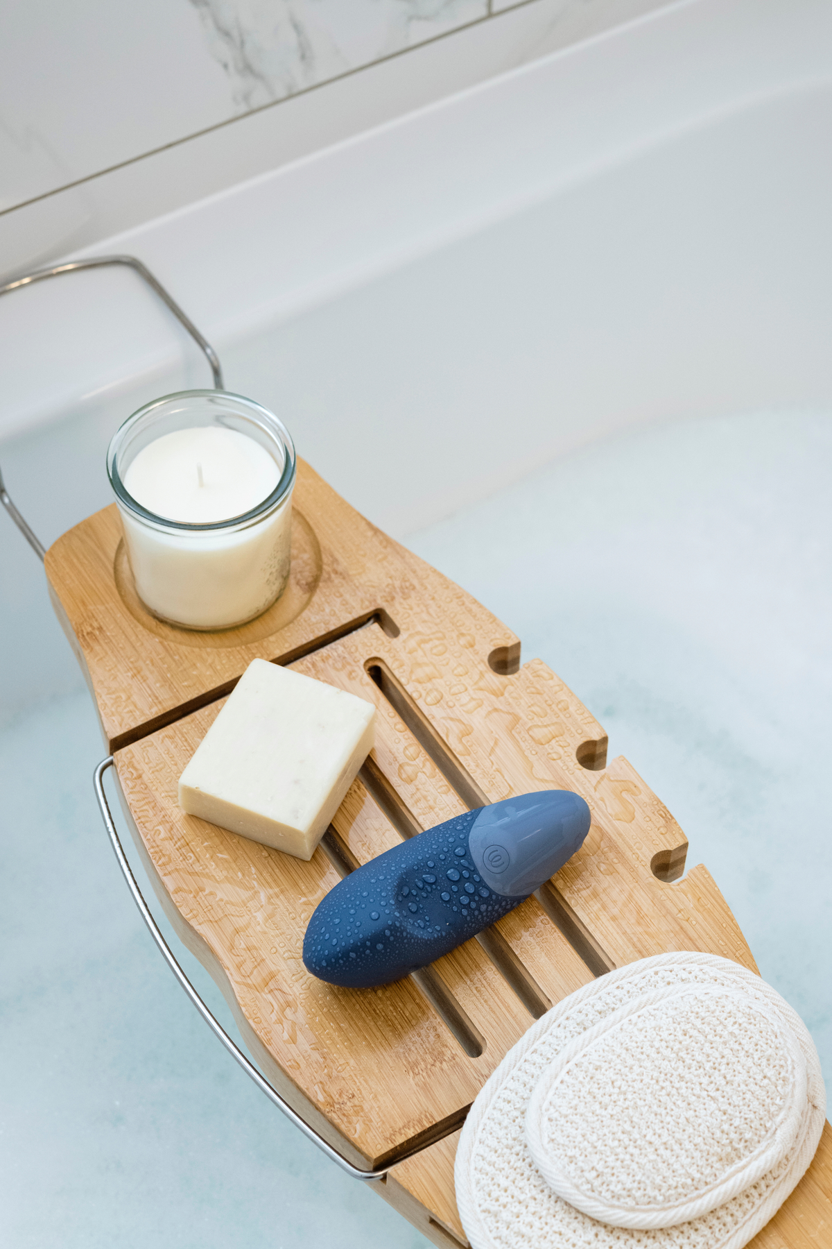 Dark blue Womanizer Vibe resting on a wooden bath caddy beside a candle, soap and sponge, showing the waterproof toy as part of a bath ritual