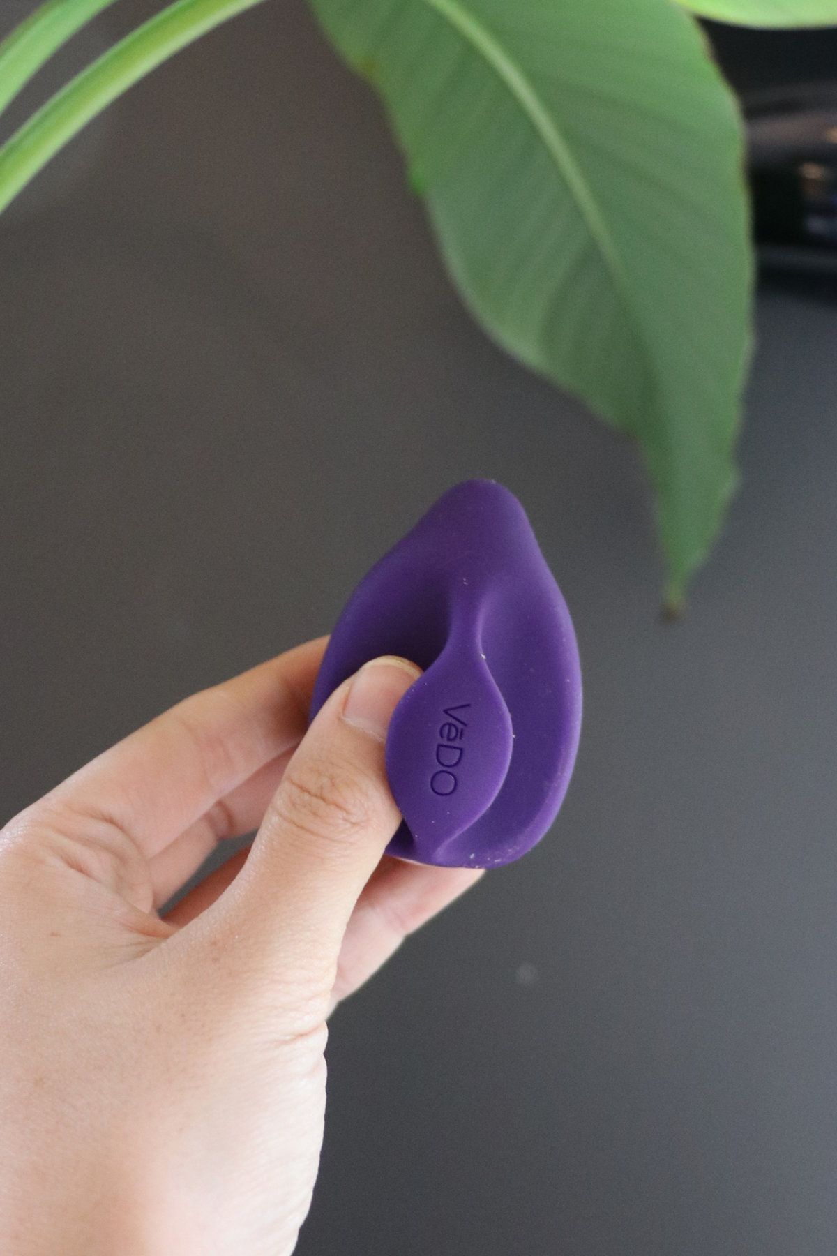 Purple Yummy Finger Vibe held between the fingers in front of a dark wall and green leaf, showing how it grips for hands-free play