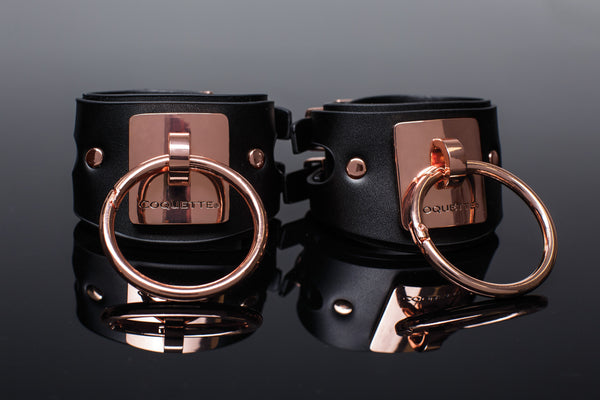 close up of the leather look texture of the vegan bondage cuffs sitting next to one another. The word  Coquette is engraved in the metal plate that holds the 2in o-ring