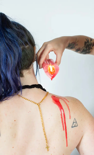 example of a lit heart shaped candle being used for wax play. red wax is dripping on white person's right shoulder; person has blue hair and faces away from camera so we see the wax on their back