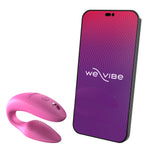 c-shaped wearable sex toy in pink next to a cell phone with a we-vibe logo on its screen. meant to show that the sex toy can be controlled via the phone app