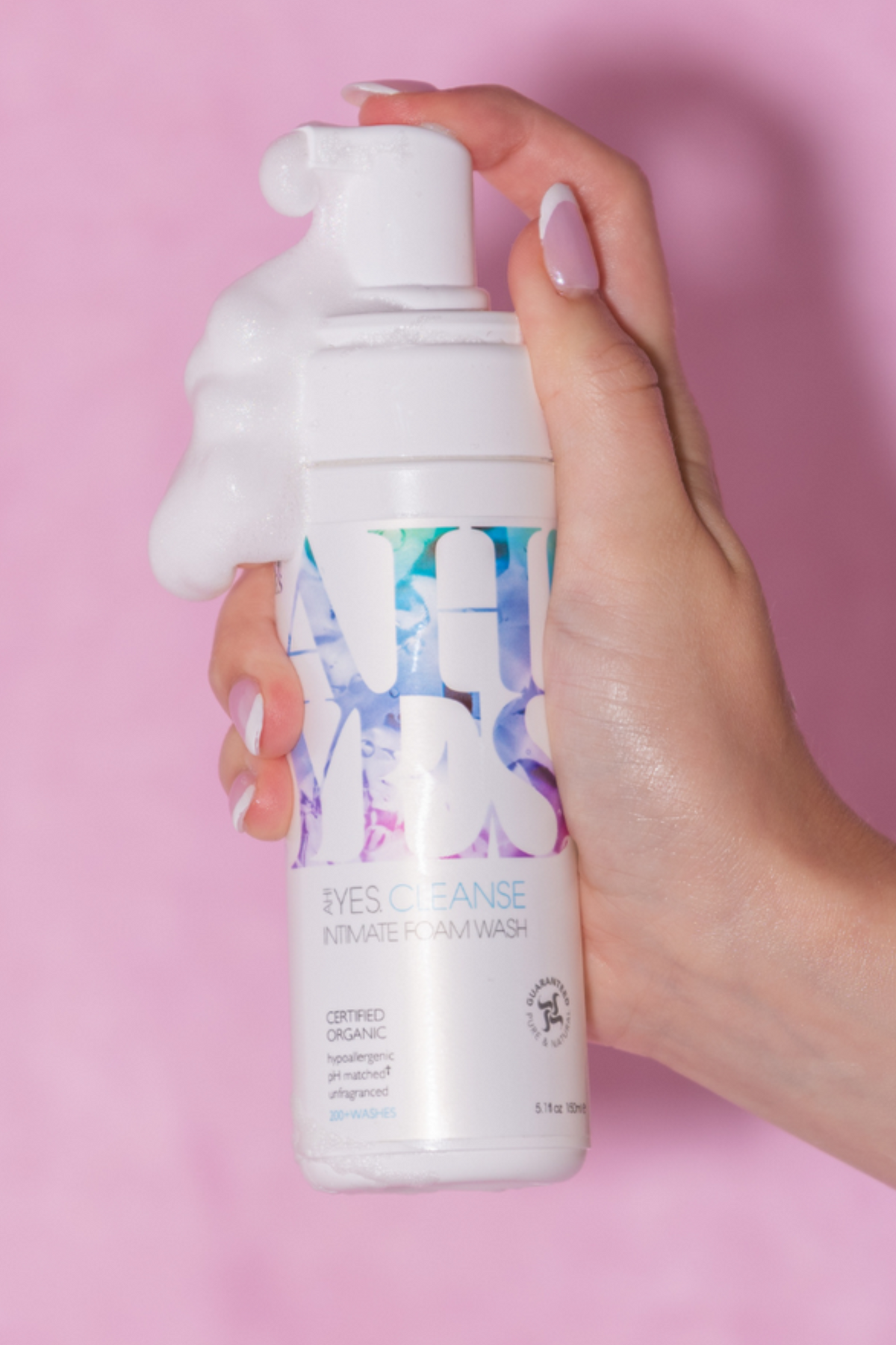 A hand holds the foaming wash bottle with a mound of white foam on the pump, held up against a pale pink background with a gentle shadow behind.