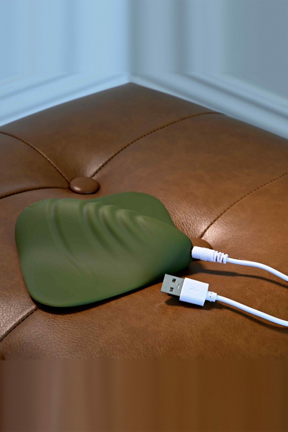 All the Pleasure grinding pad in Olive Green on a leather ottoman with its USB charging cable plugged in at the side