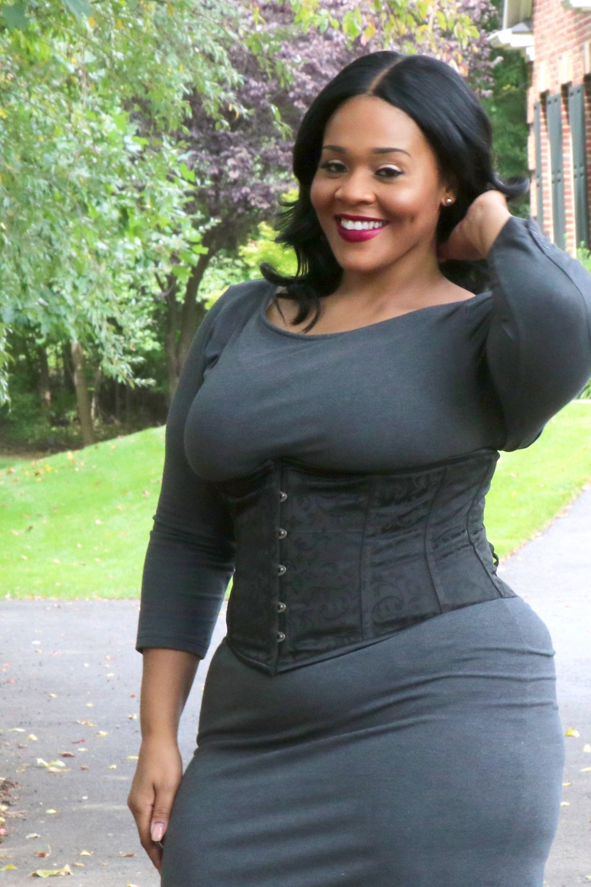 Smiling plus-size model outdoors wearing the black brocade Alluring underbust corset over a fitted grey long-sleeve dress