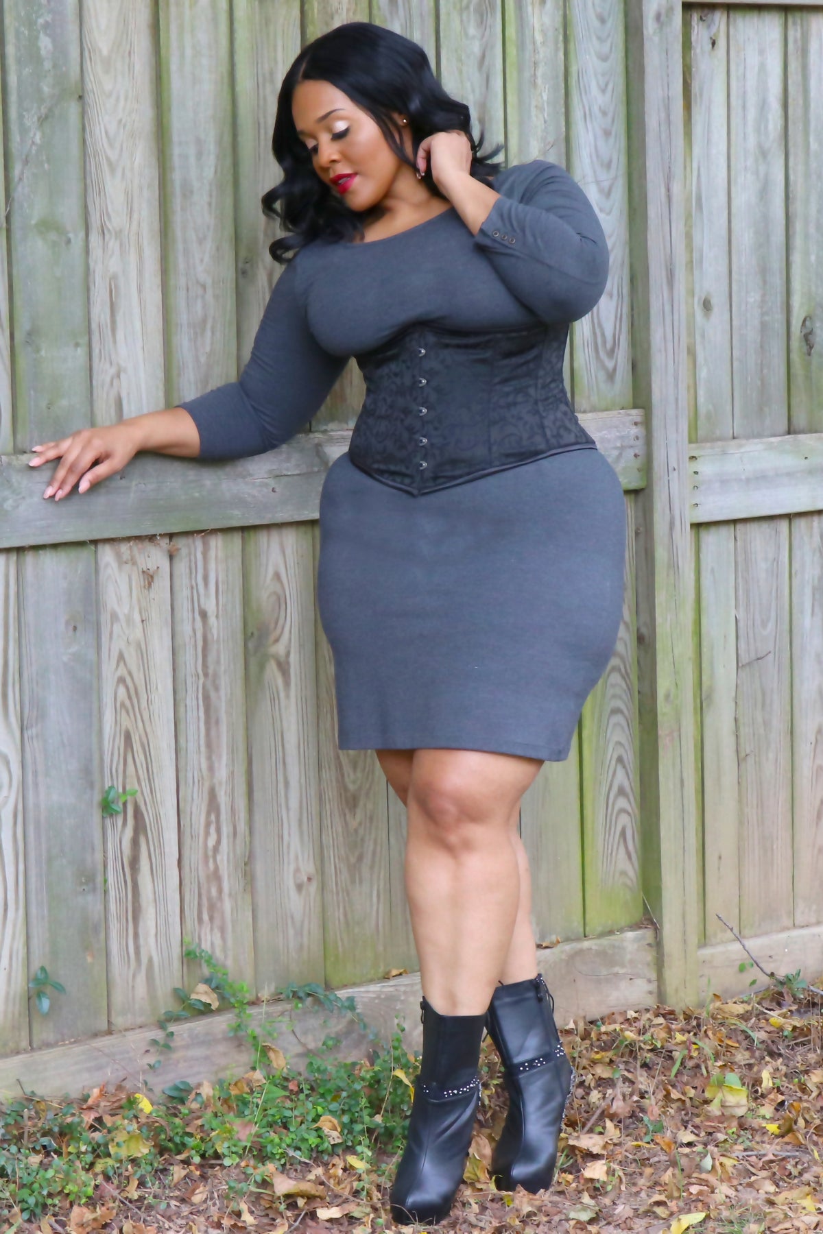 Plus-size model posing against a wooden fence in the Alluring black brocade underbust corset, grey dress and black ankle boots