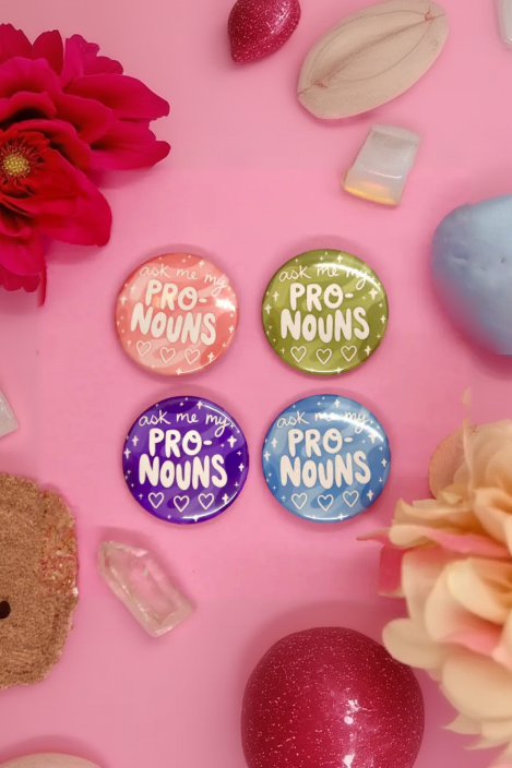 Four Ask Me My Pronouns pinback buttons in peach, green, purple and blue on a pink surface with flowers and crystals
