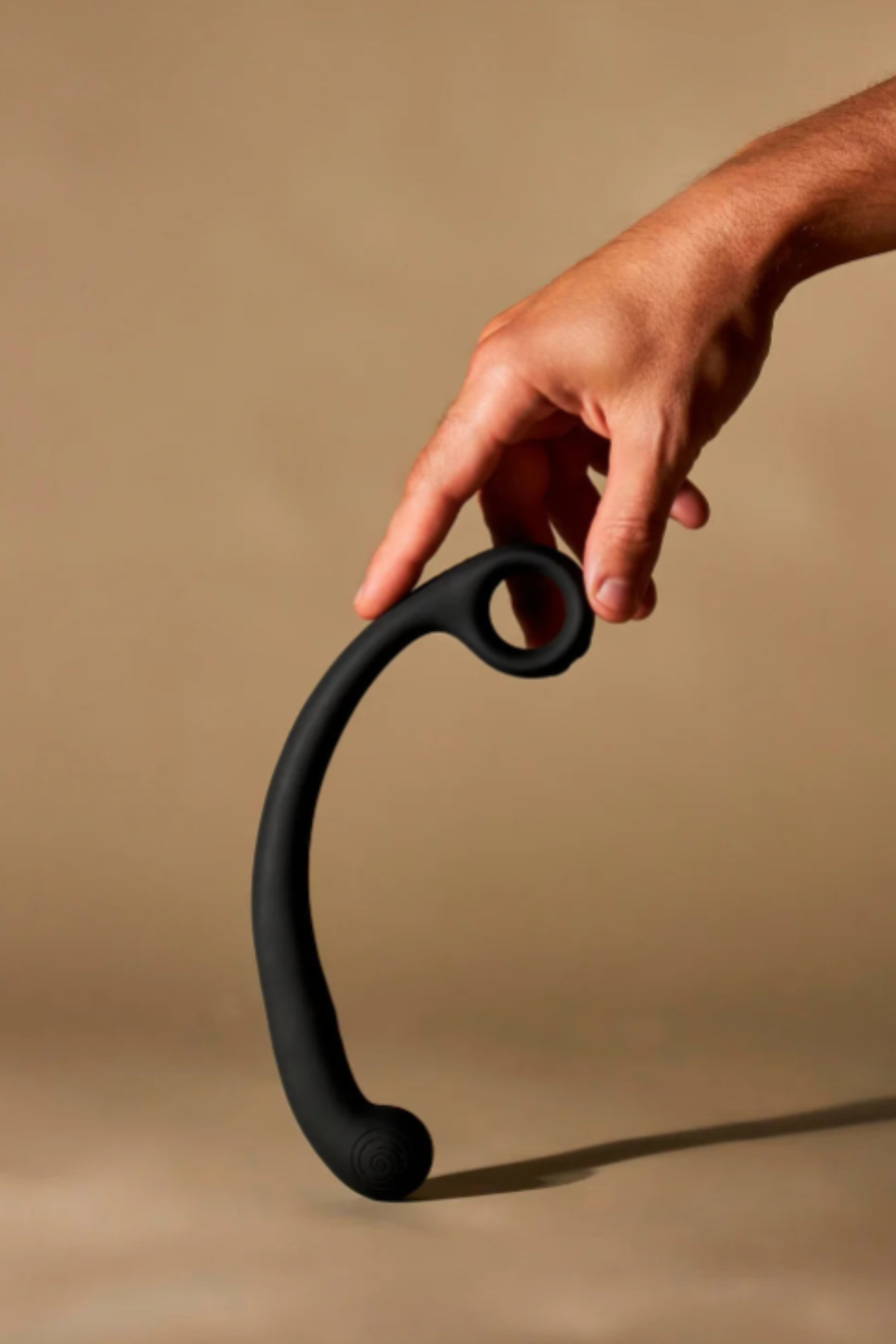 Hand lifting the Granite Noir Axi Slimline G-spot wand by its finger loop against a warm beige backdrop, showing its slim curved profile