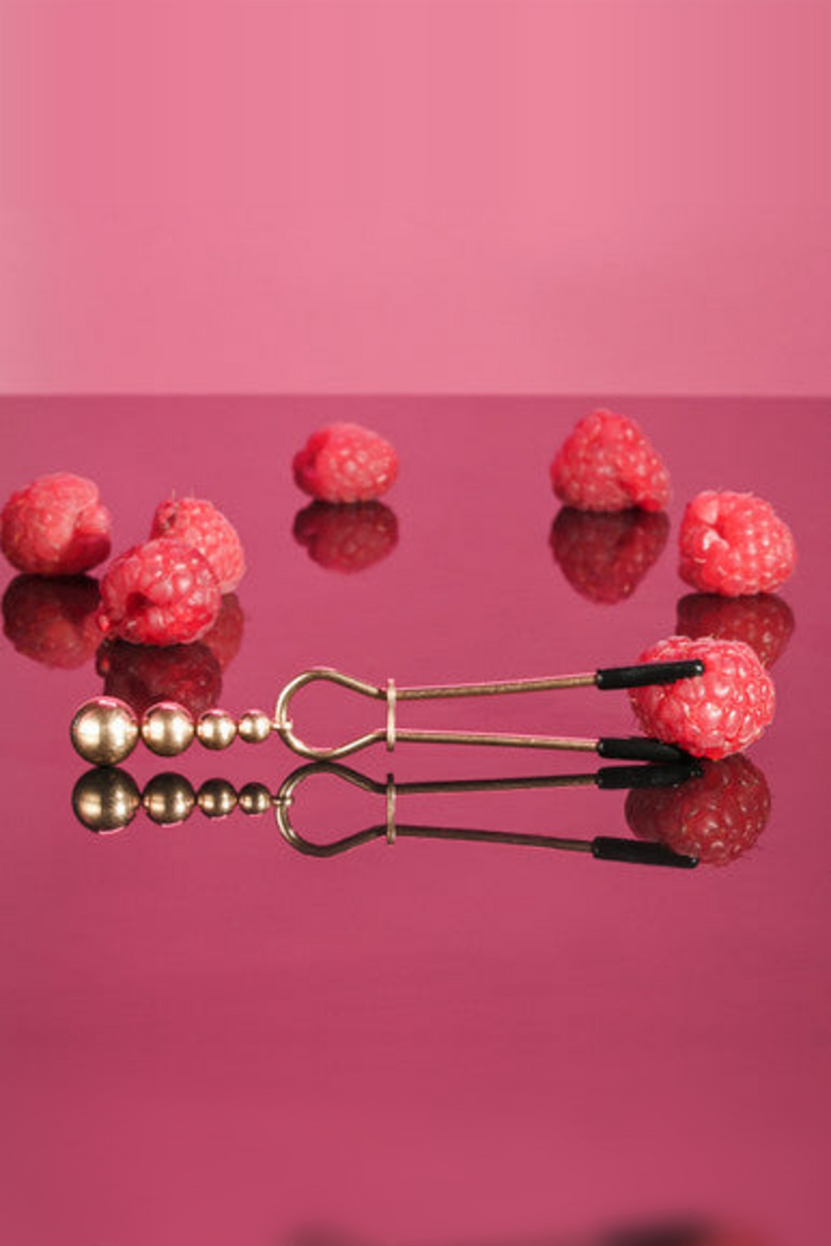 Rose gold beaded tweezer nipple clamp lying on a glossy pink surface among raspberries, showing its slide ring and ball chain