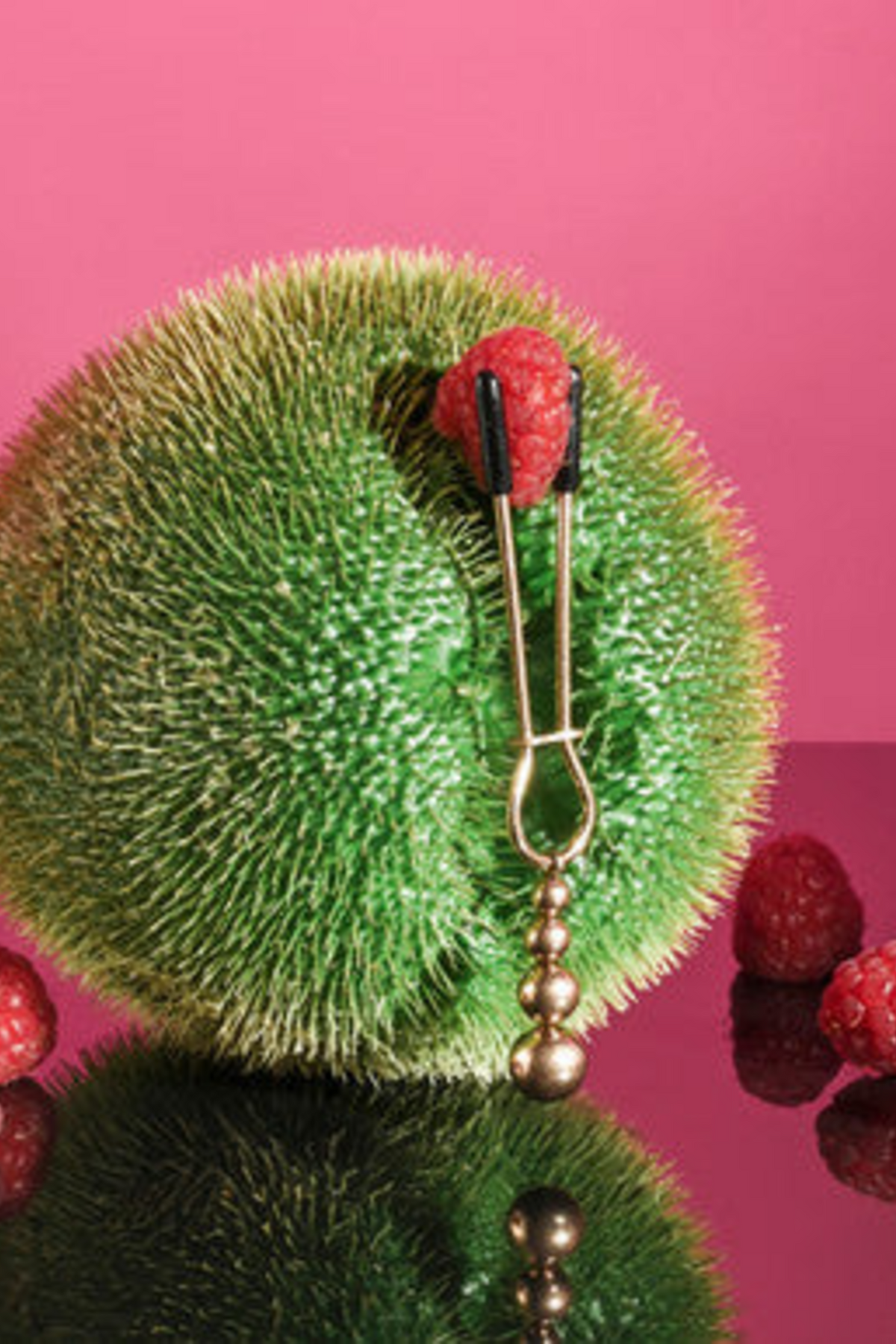 Rose gold tweezer nipple clamp gripping a raspberry on a green spiky ball, showing the slide ring and ball weight on pink