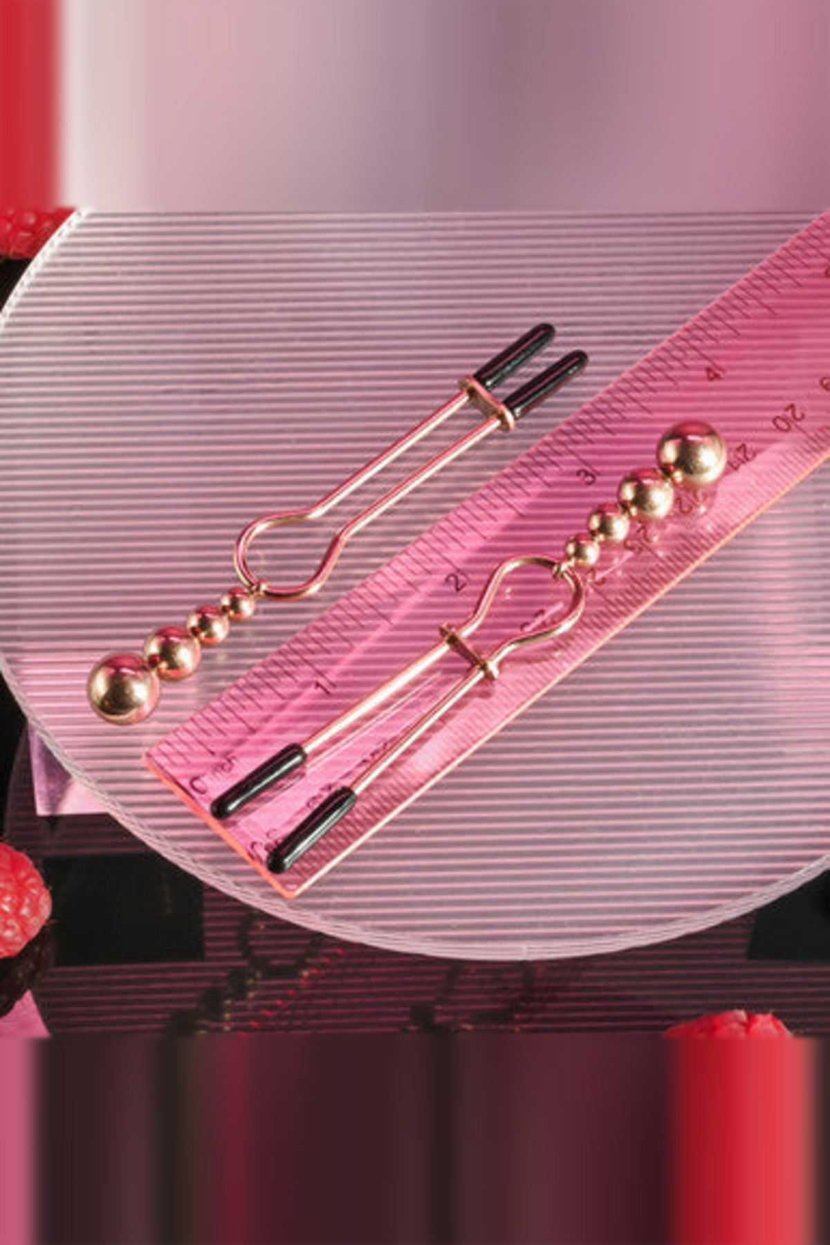 Pair of rose gold beaded nipple clamps lying on a pink ruler, showing their full length, slide rings and rubber-tipped jaws