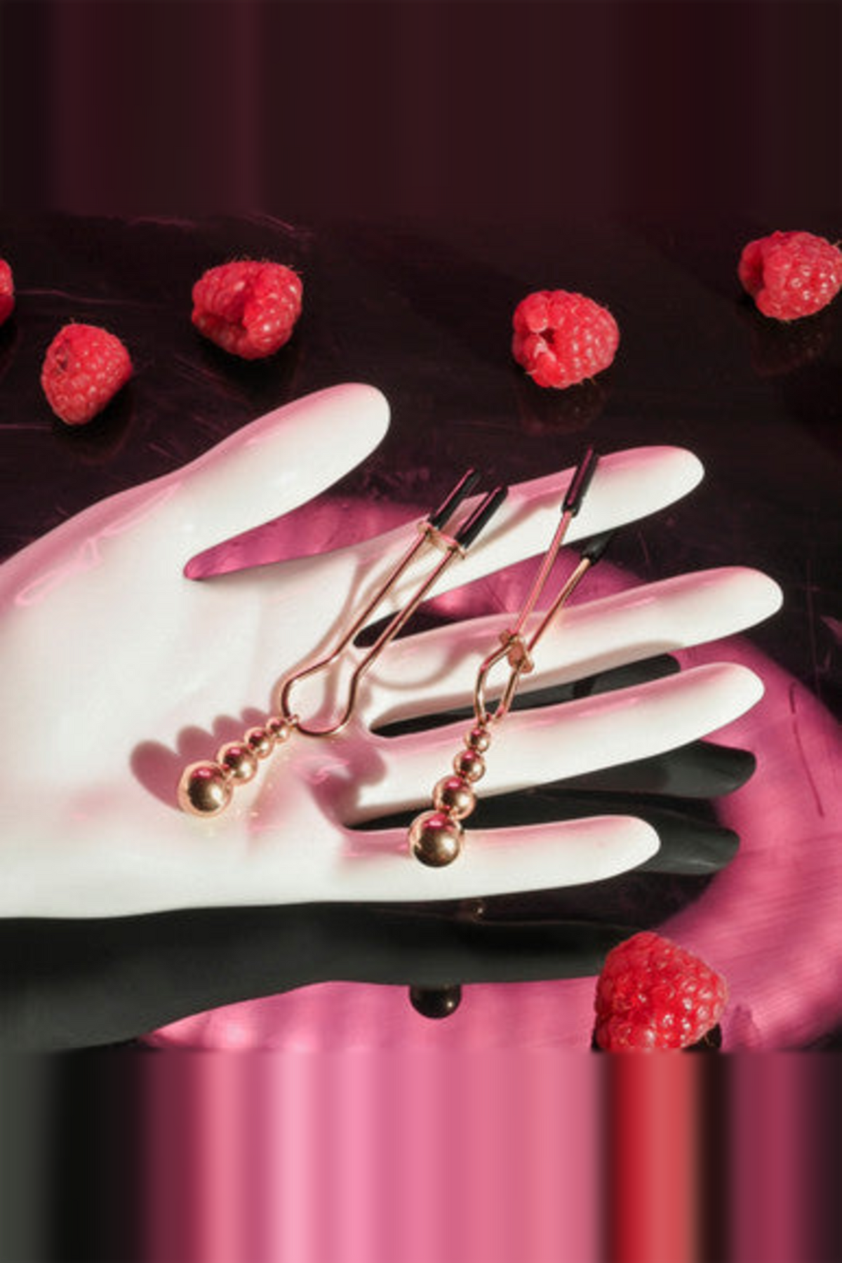 Rose gold beaded nipple clamps resting on a white rubber glove with raspberries, showing their slim tweezer shape and weights