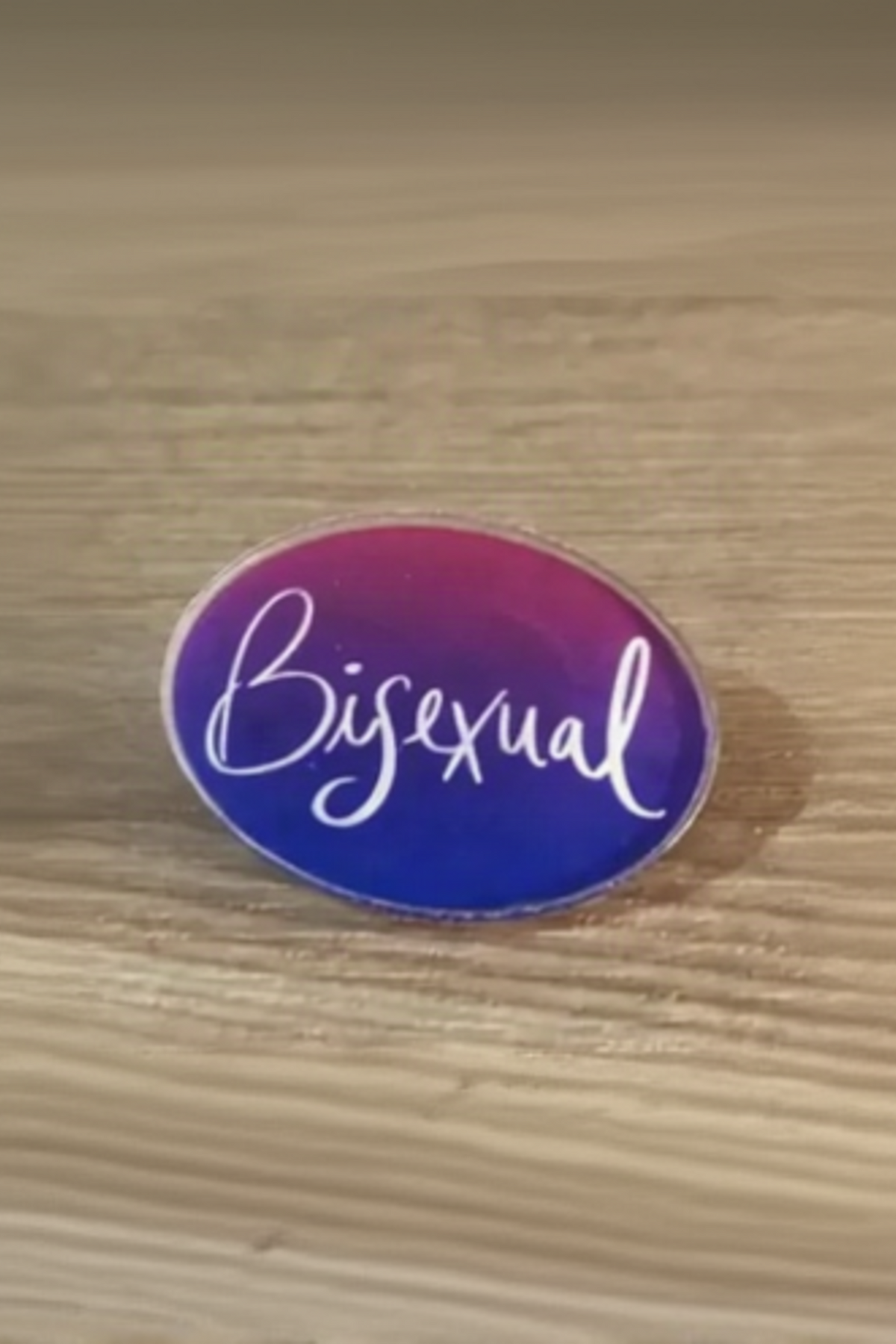 Oval acrylic pin with a pink-to-purple-to-blue bisexual flag gradient and the word Bisexual in white script, resting on a light wood surface.