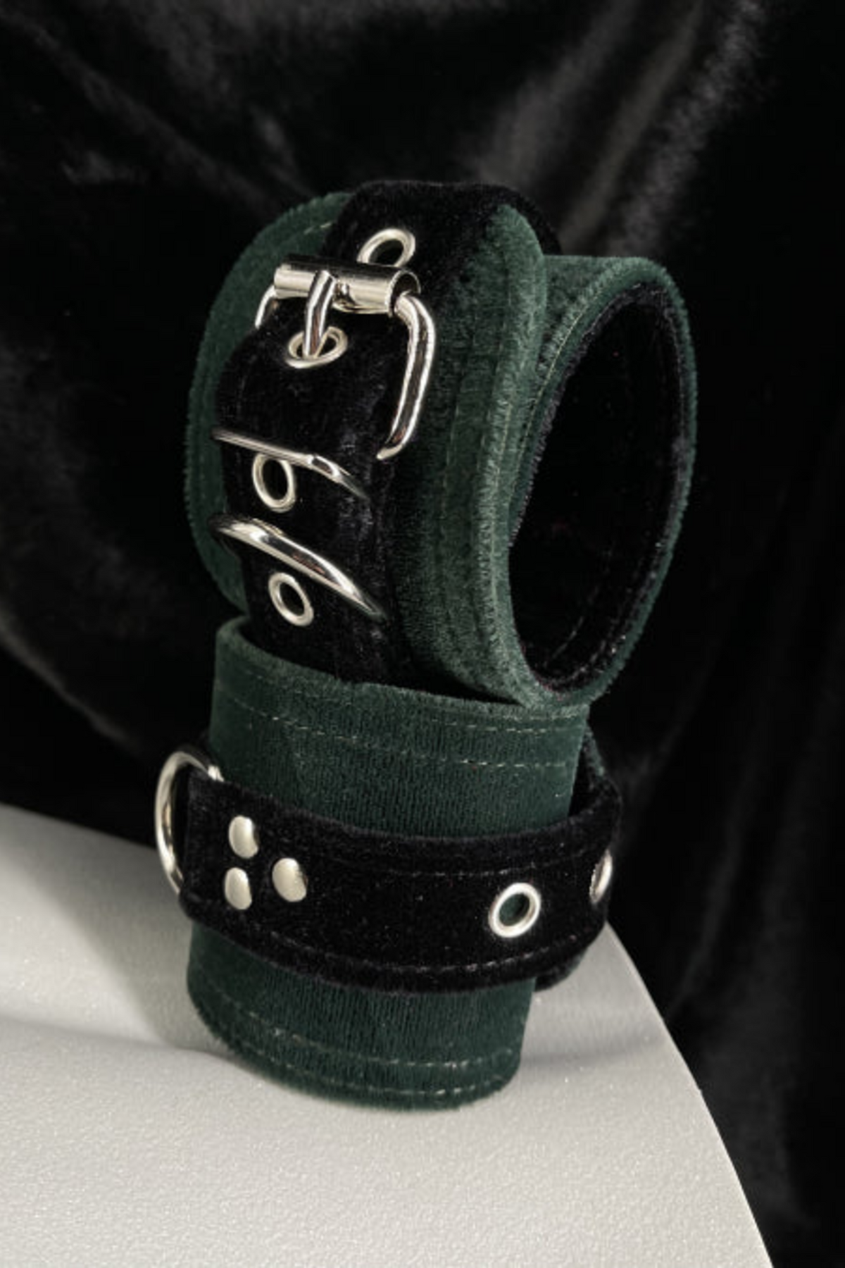 Green and black velvet D-ring cuffs with silver buckles stacked on a white mannequin form, set against a backdrop of draped black velvet fabric.