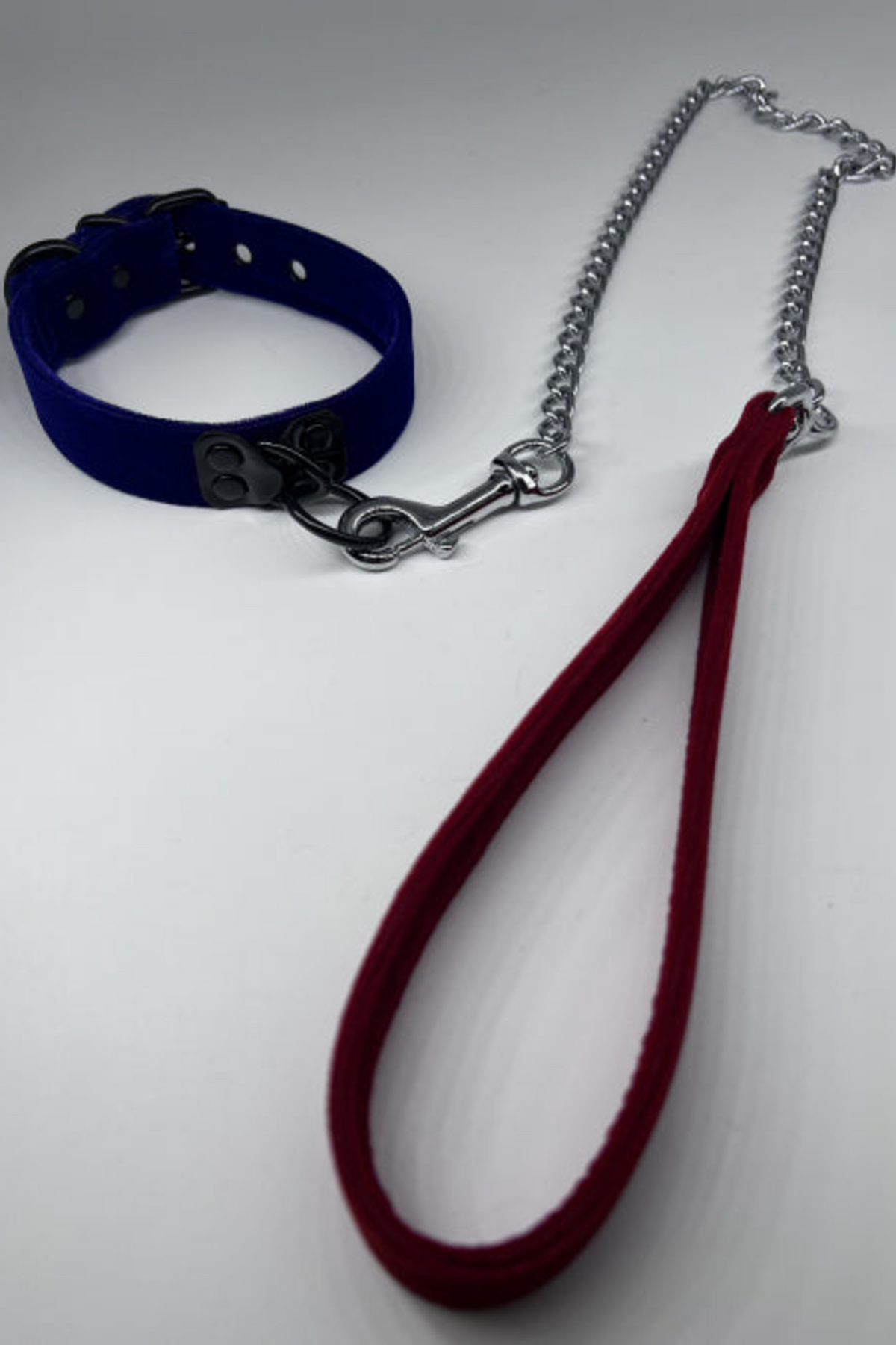 Burgundy velvet leash with its silver chain clipped by the snap hook to a navy velvet collar, the velvet hand loop lying in front on a pale grey backdrop.