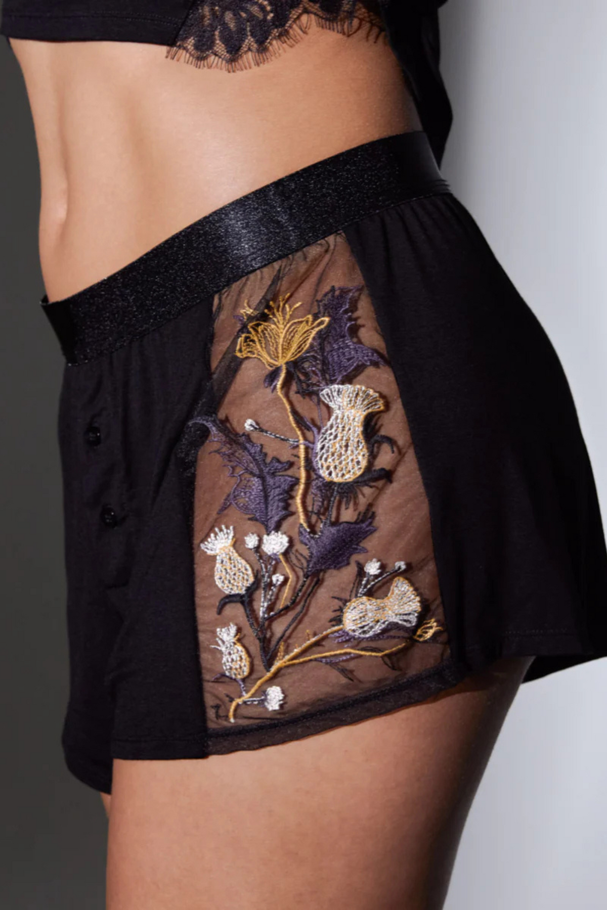 Side close-up of the black Cirsi the Sorceress modal shorts with a glossy elastic waistband and sheer side panel of embroidered thistles