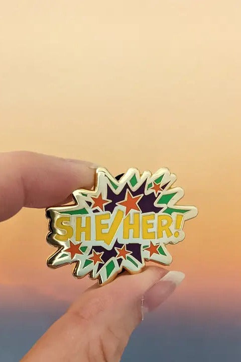 Fingers holding the Comic POW! She/Her enamel pin, a gold-edged starburst with yellow lettering and colourful stars, against a sunset sky