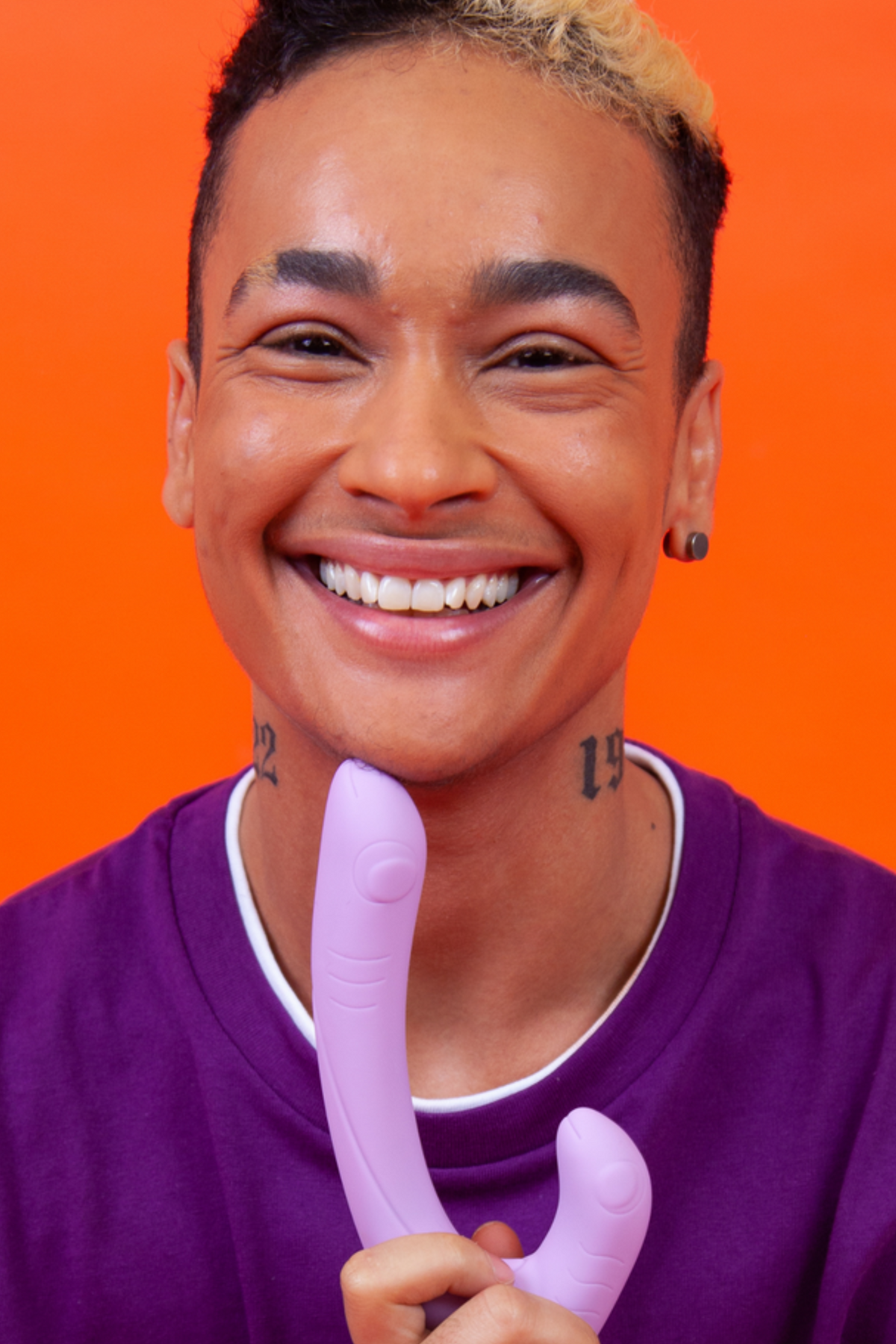 Smiling person in a purple top holding the lilac Cute Little Fuckers Jix slim rabbit vibrator in front of a bright orange background