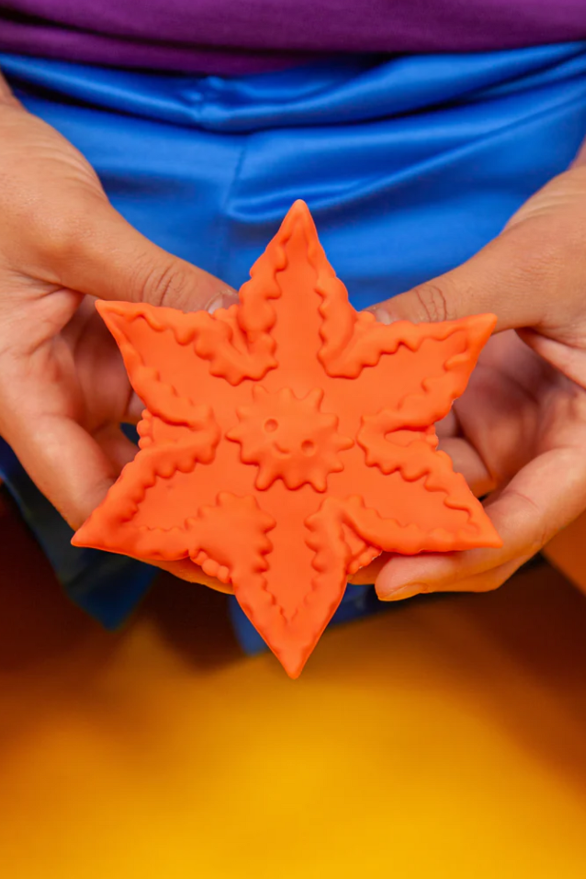 Hands holding the coral Cute Little Fuckers Starsi star shaped vibrator in front of a blue top on a bright yellow background