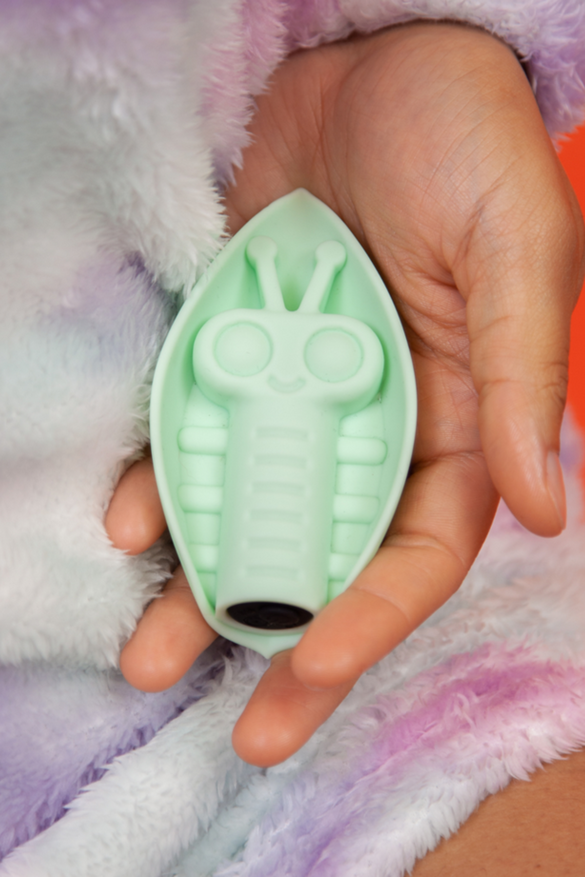 Mint Zeep grinding vibe resting in an open palm against a fluffy pastel tie-dye robe, its smiling bug-faced leaf pad facing up and charge port showing.