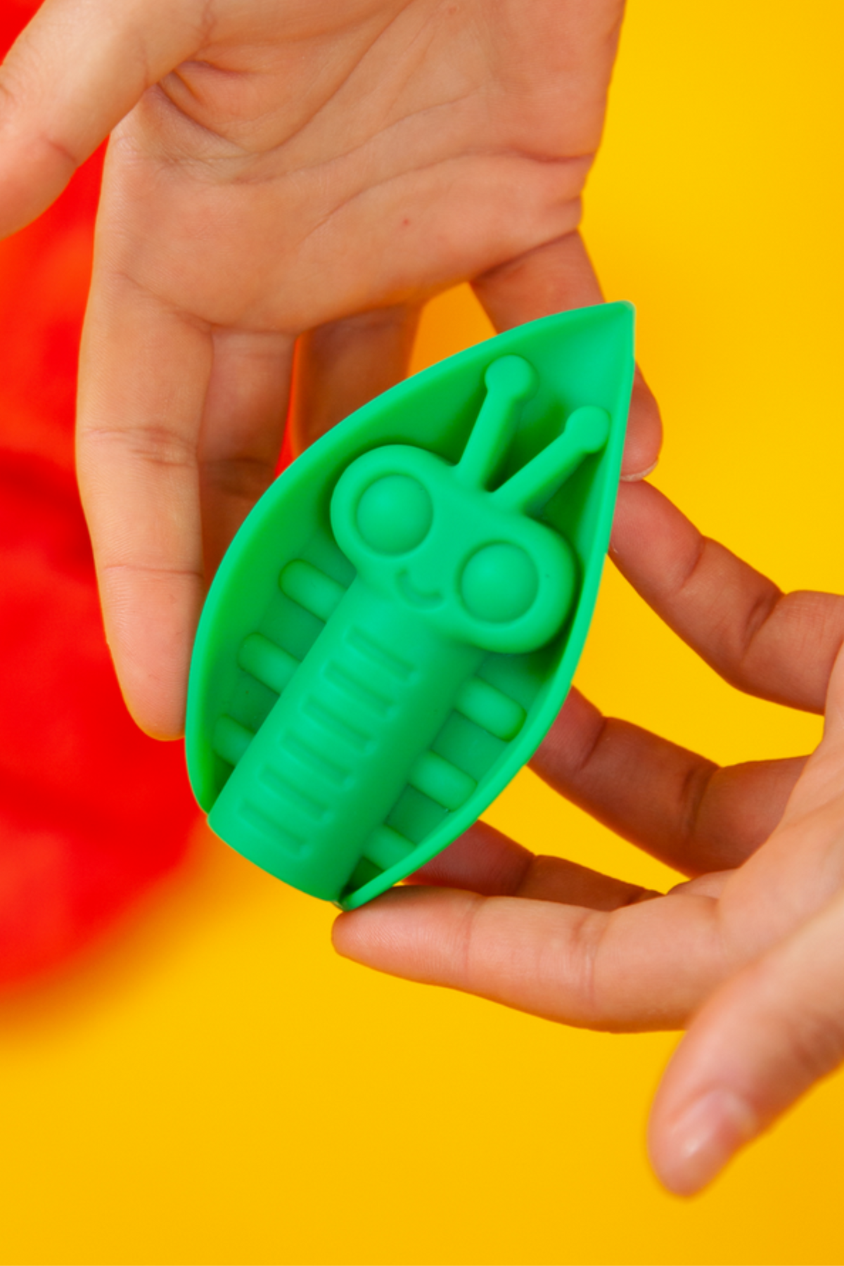 Two hands holding the emerald Zeep grinding vibe against bold red and yellow, the leaf-shaped pad tilted to show its smiling raised bug design.