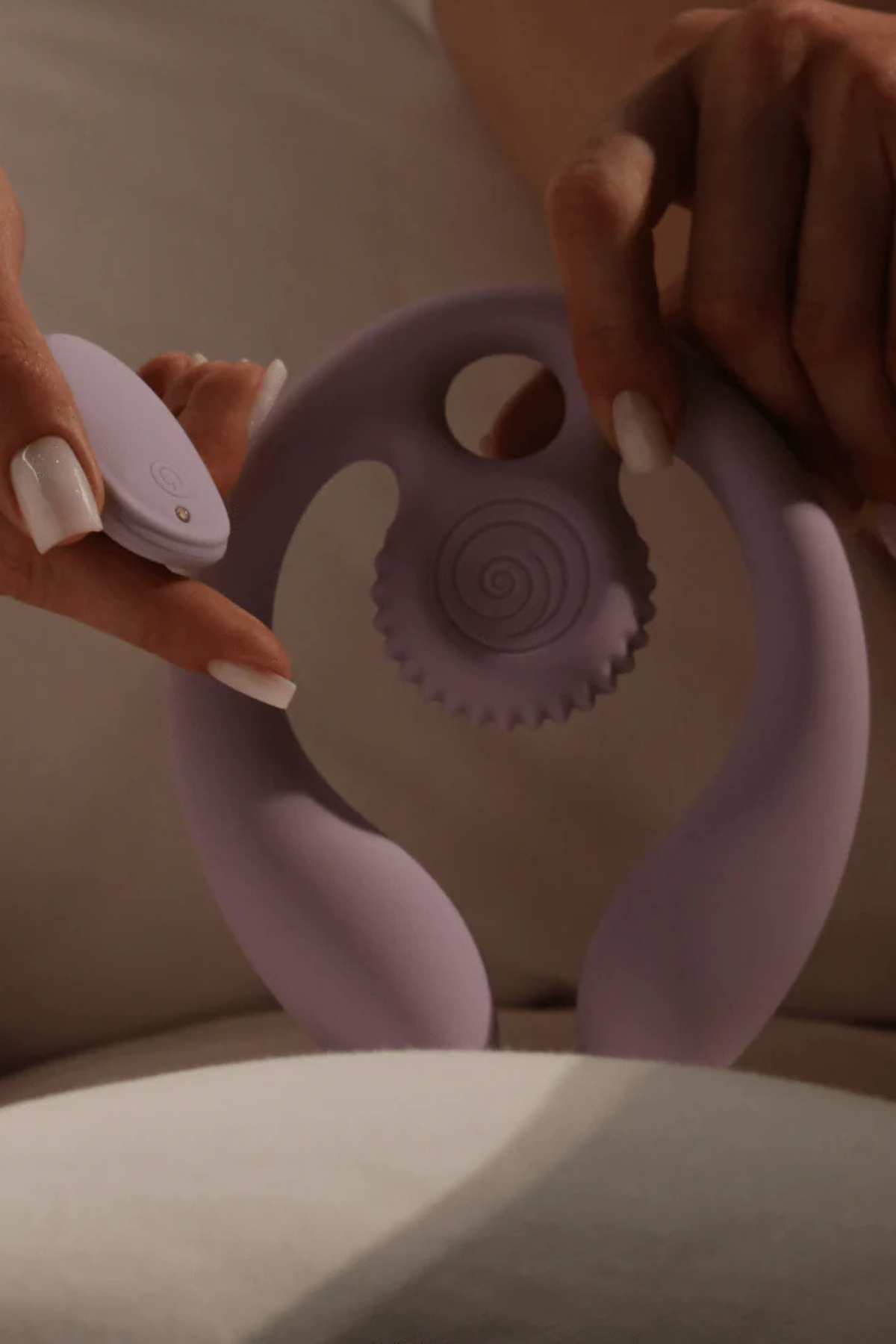 Hands holding the lilac Gizi Duo over a cream sofa, its remote in one hand, showing the flexible arms and textured snail head up close