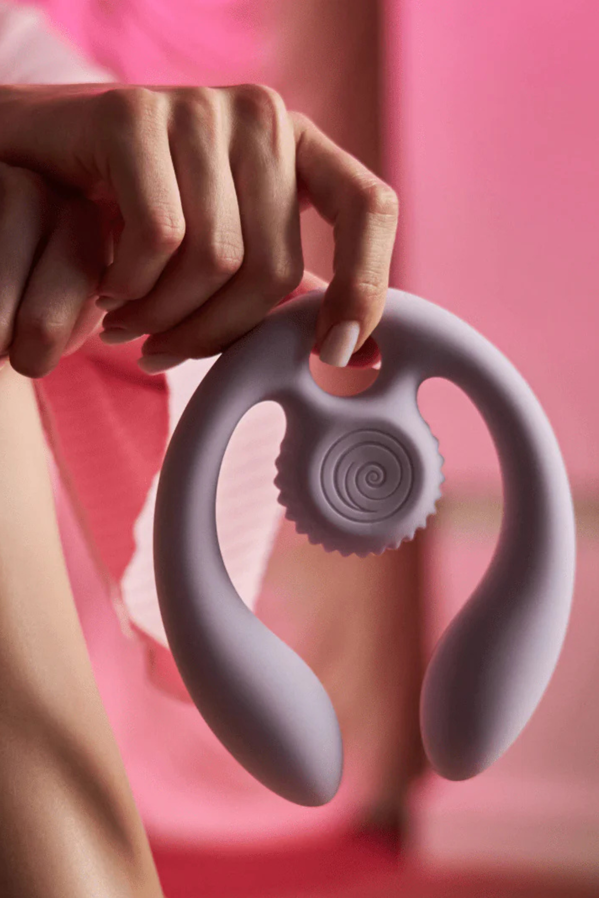 Hand holding the lilac Gizi Duo by its finger loop against a bright pink backdrop, letting the dual-ended vibrator hang to show its shape