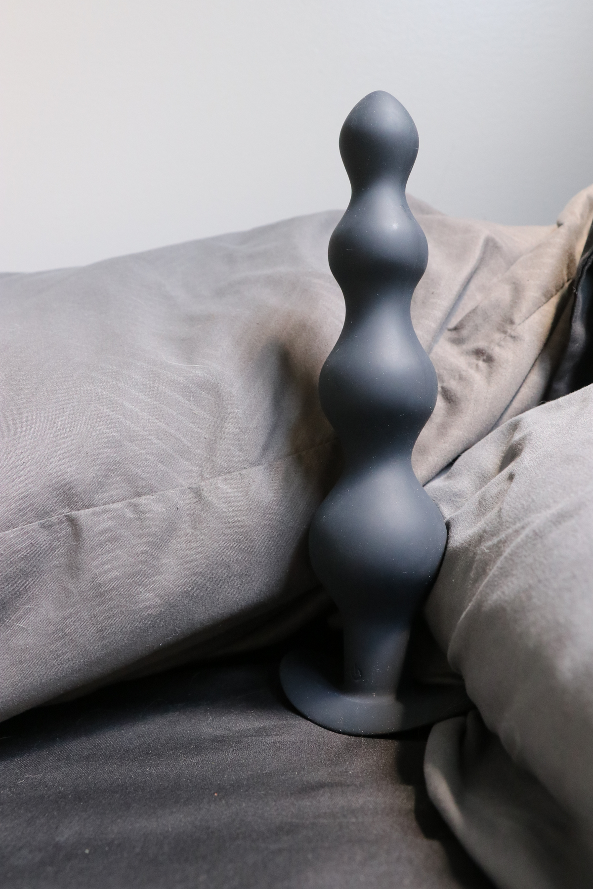 The black beaded wand standing upright on a grey bed with soft rumpled bedding and a pillow behind it, lit by daylight from the side.