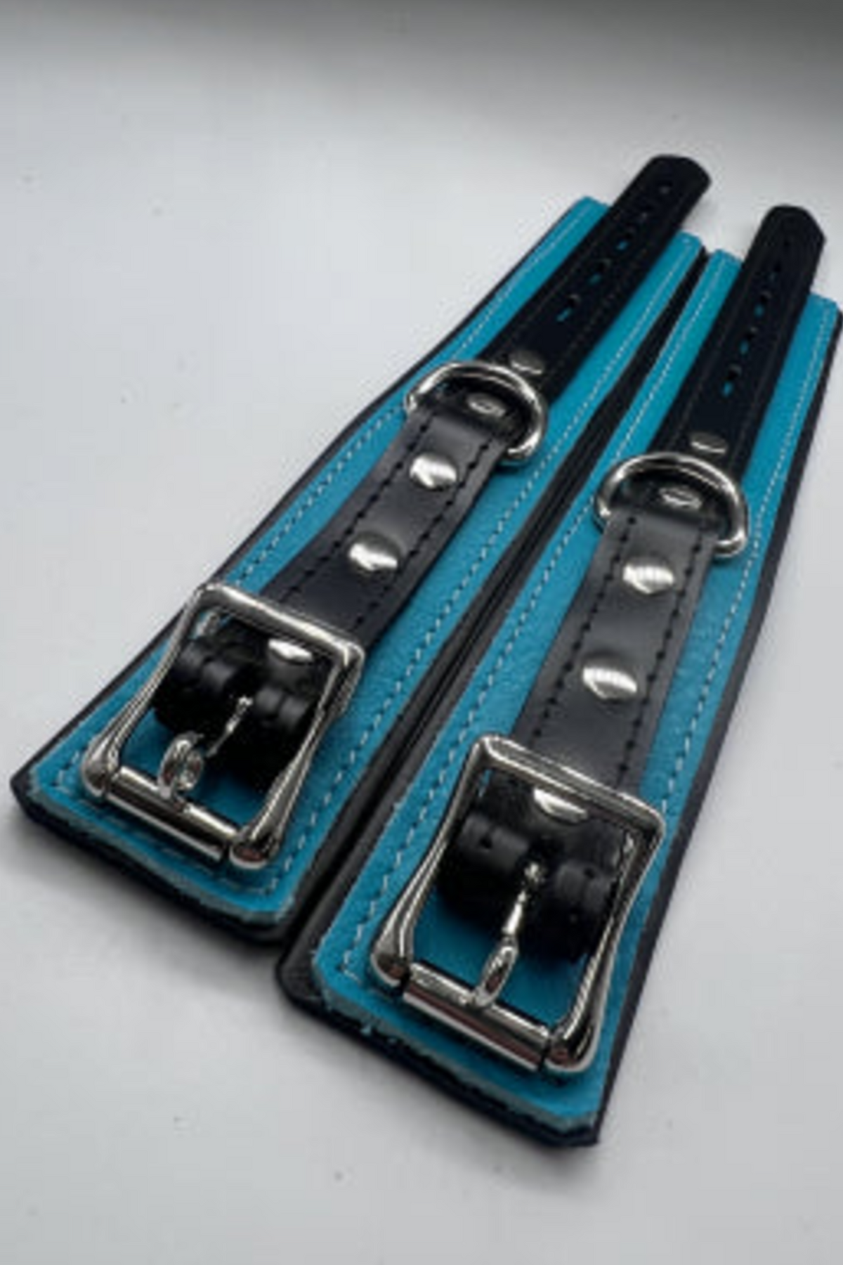 Two teal and black leather locking cuffs laid flat side by side on a white surface, showing their silver roller buckles, D-rings, rivets and adjustment holes.