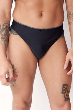Close front view of the Floruisse Lucy lace thong worn by a standing model: smooth black gaff front, bow detail, sage lace sides