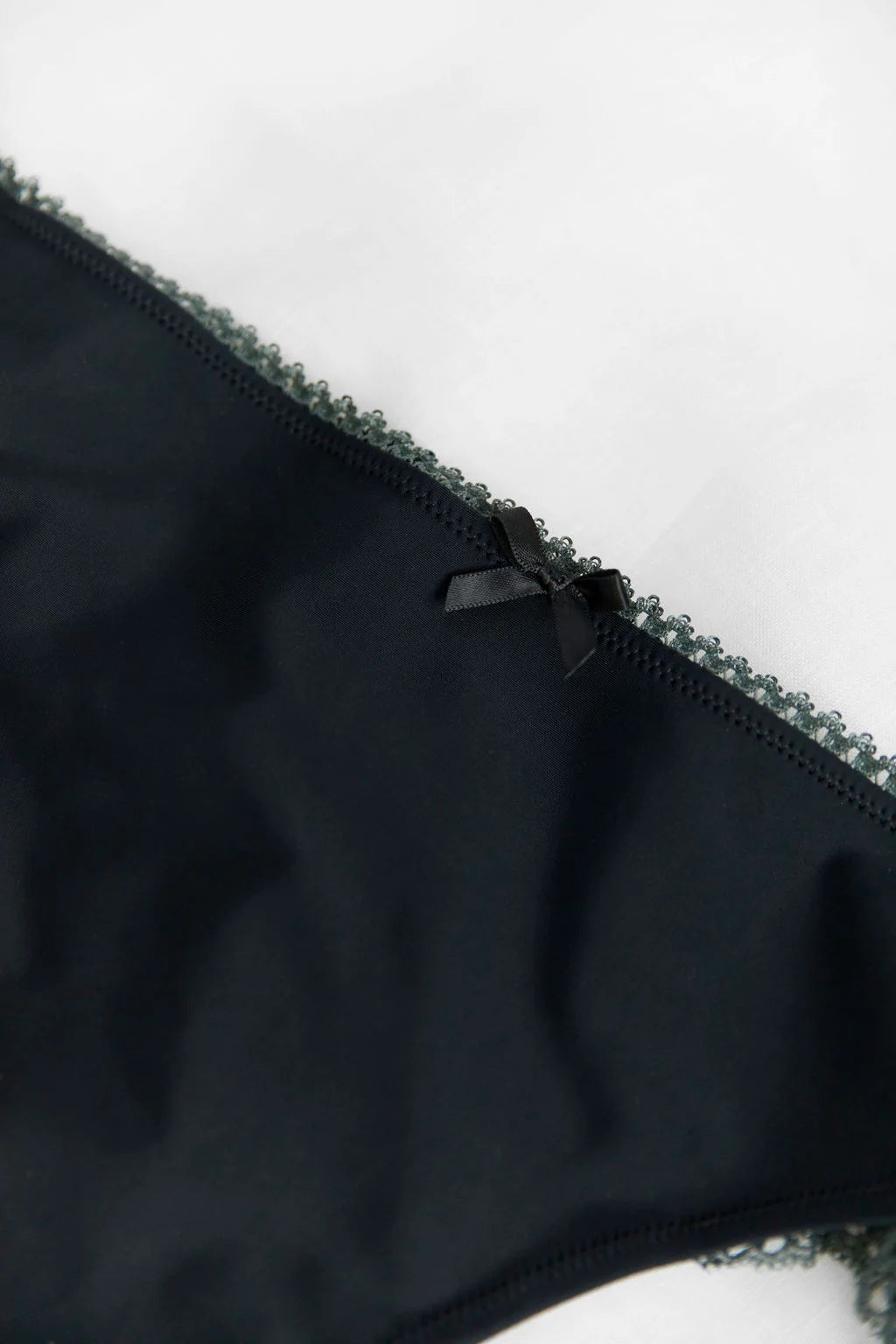 Close-up of the Floruisse Lucy thong waistband showing the black jersey fabric, small bow detail and fine picot edge trim