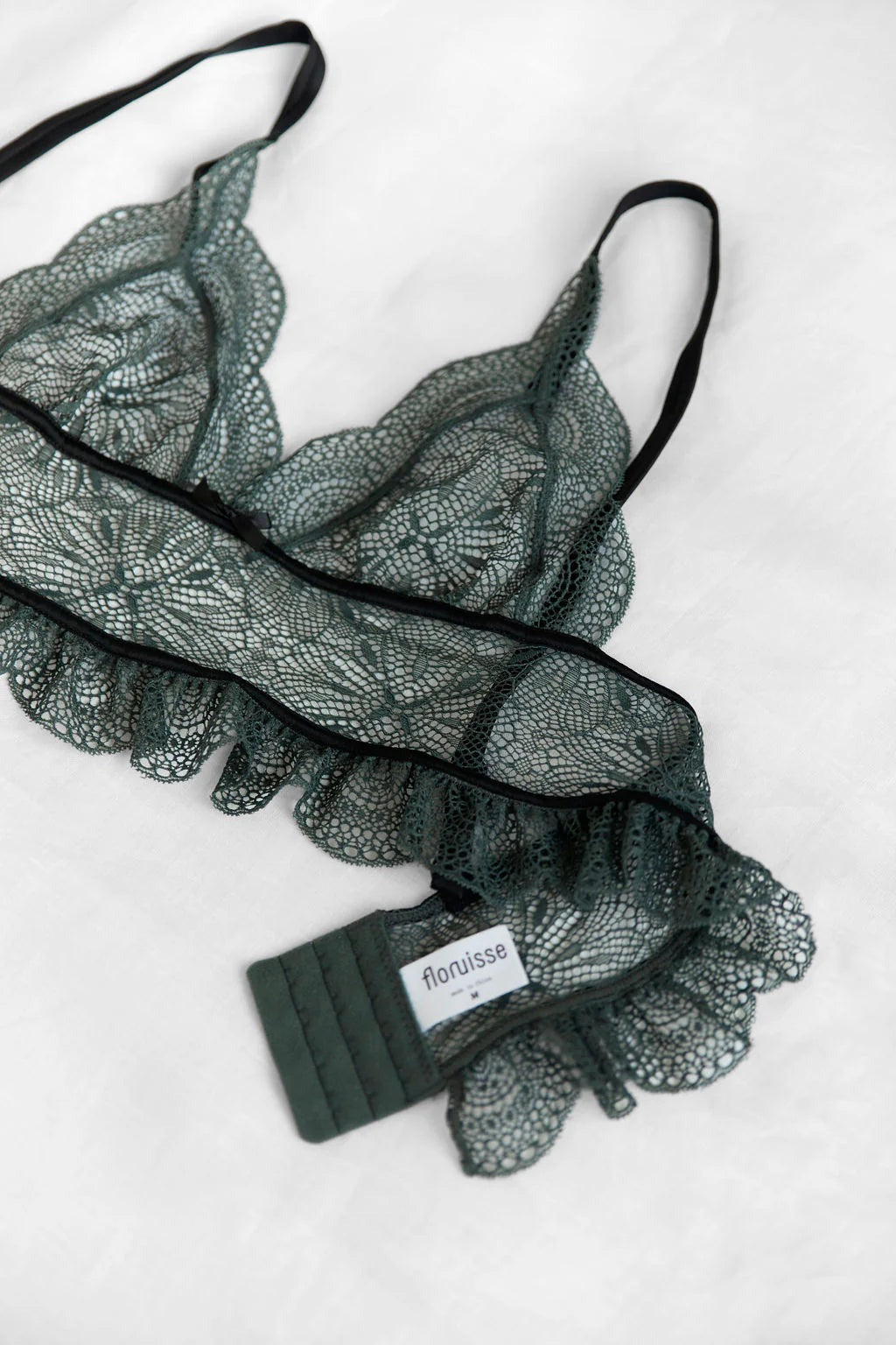 Close-up of the Floruisse Ru bralette in slate sage lace on white, showing the scalloped edges, black straps and brand label