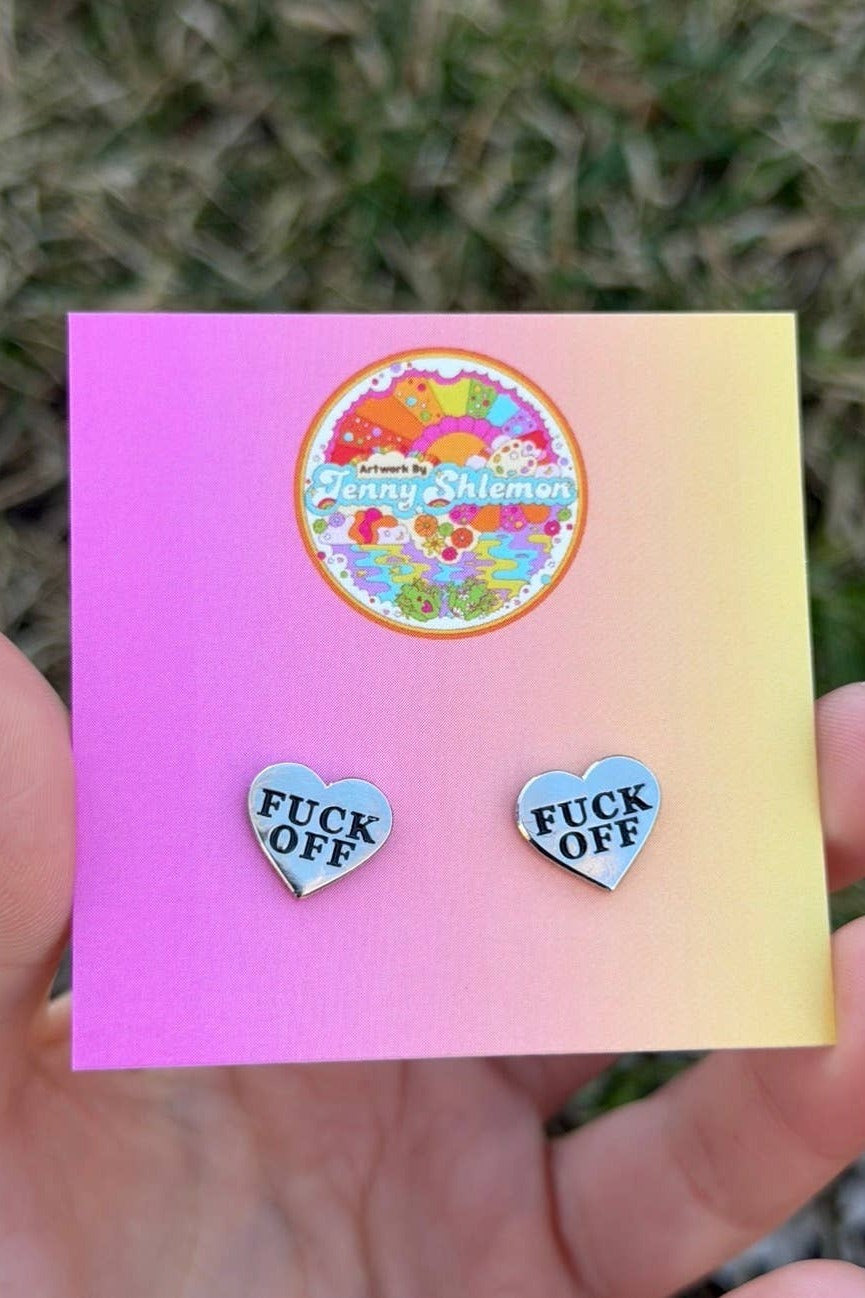 Hand holding a pink-to-yellow backing card with a pair of silver enamel heart stud earrings reading Fuck Off, photographed over grass