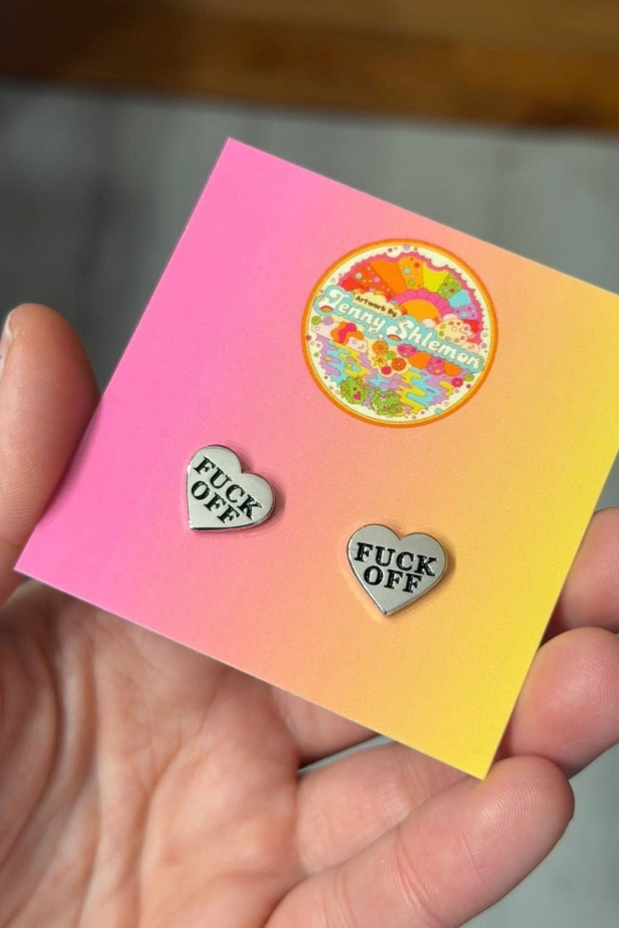 Angled close view of silver enamel Fuck Off heart stud earrings on a pink and yellow gradient ShlemonArt backing card held in a palm