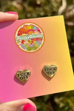 Gold enamel heart stud earrings reading Fuck Off on a pink and yellow backing card, held by fingers with pink nail polish over grass