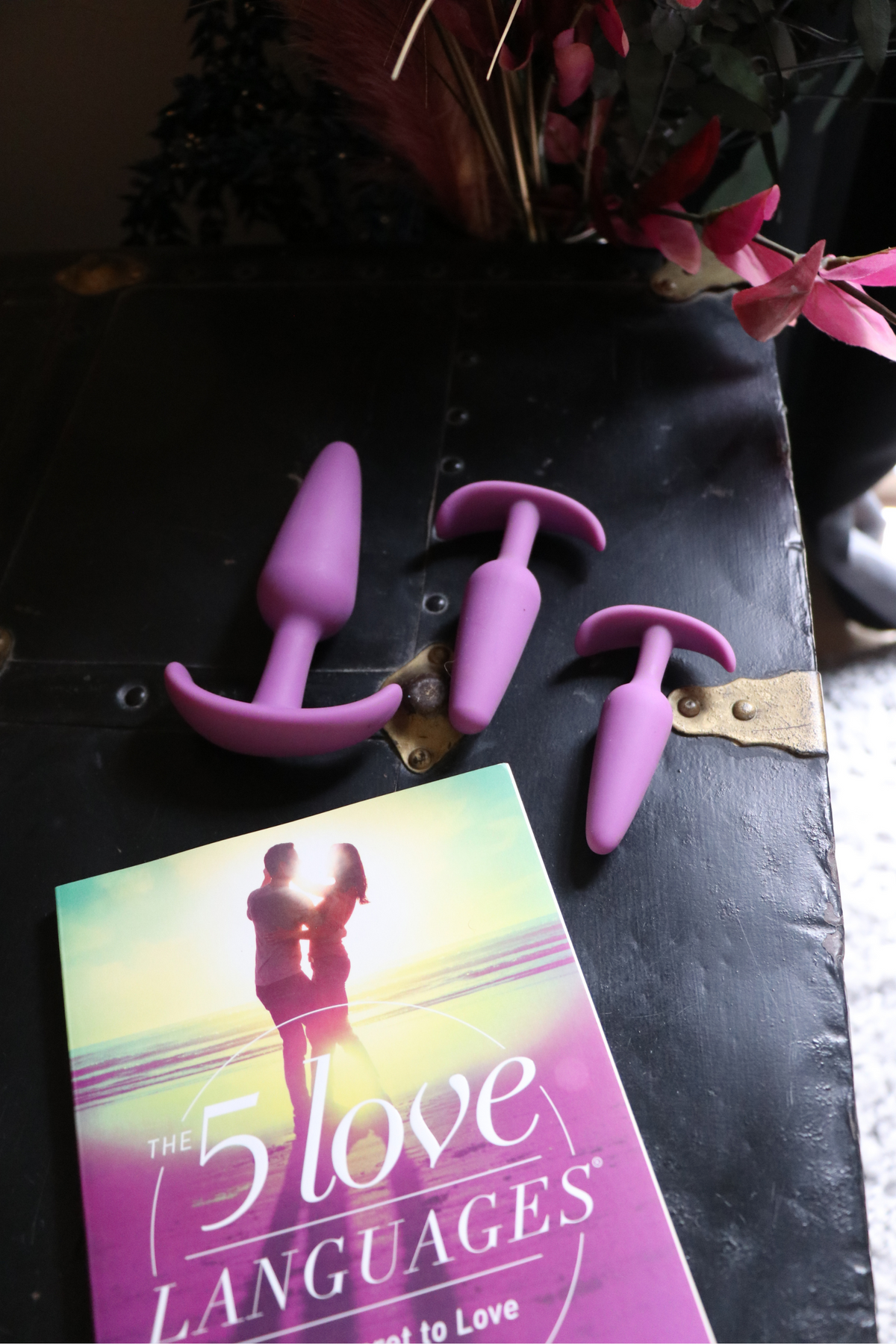The three purple Gossip plugs arranged on a black leather surface beside red flowers and a copy of The 5 Love Languages book.