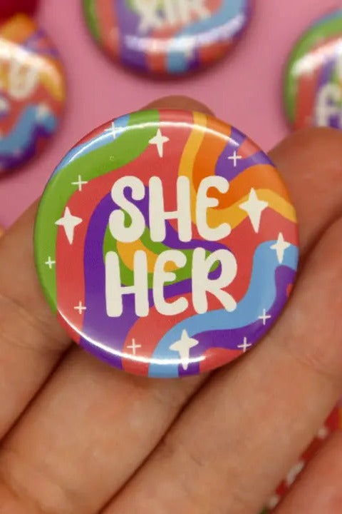Hand holding a 1.5 inch holographic pronoun button reading She Her in white lettering on a wavy rainbow background with sparkles