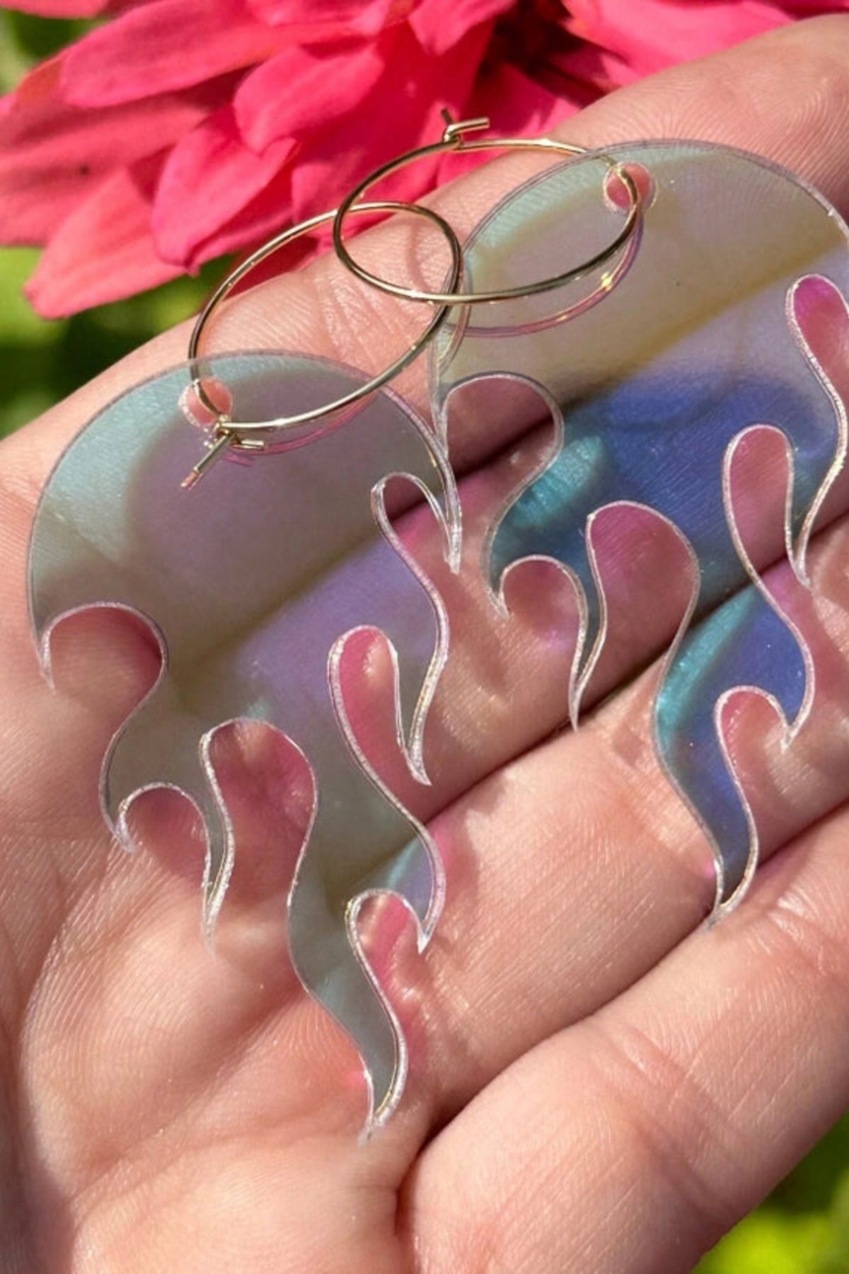 The same flame hoop earrings in a pale pink and lilac iridescent finish, held on an open palm outdoors with a bright pink flower behind.