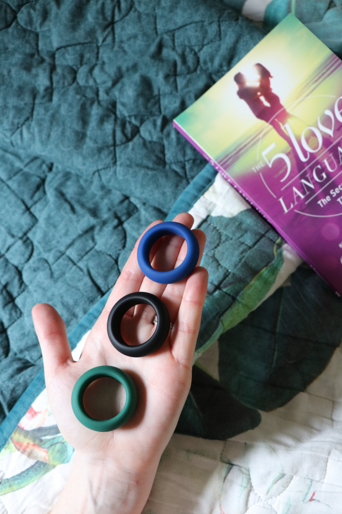 A hand holding the three silicone rings above a teal bedspread, with a book propped behind, showing the black, green and blue sizes.