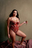 plus size light skinned black model stands posing with one foot up on a step holding out a gold challis. She is wearing the Kilo Brava Embroidery Teddy in Ruby Wine