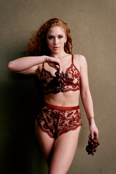 curly redheaded model in size small wears embroidery longling bralette and matching tap shorts in ruby wine red color. model is posing with grapes