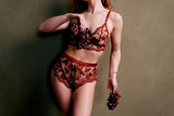 shows the torso of a skinny white person with red hair wearing a beautiful embroidered matching lingerie set in deep ruby wine red color. they are holding grapes artfully posed in front of their chest