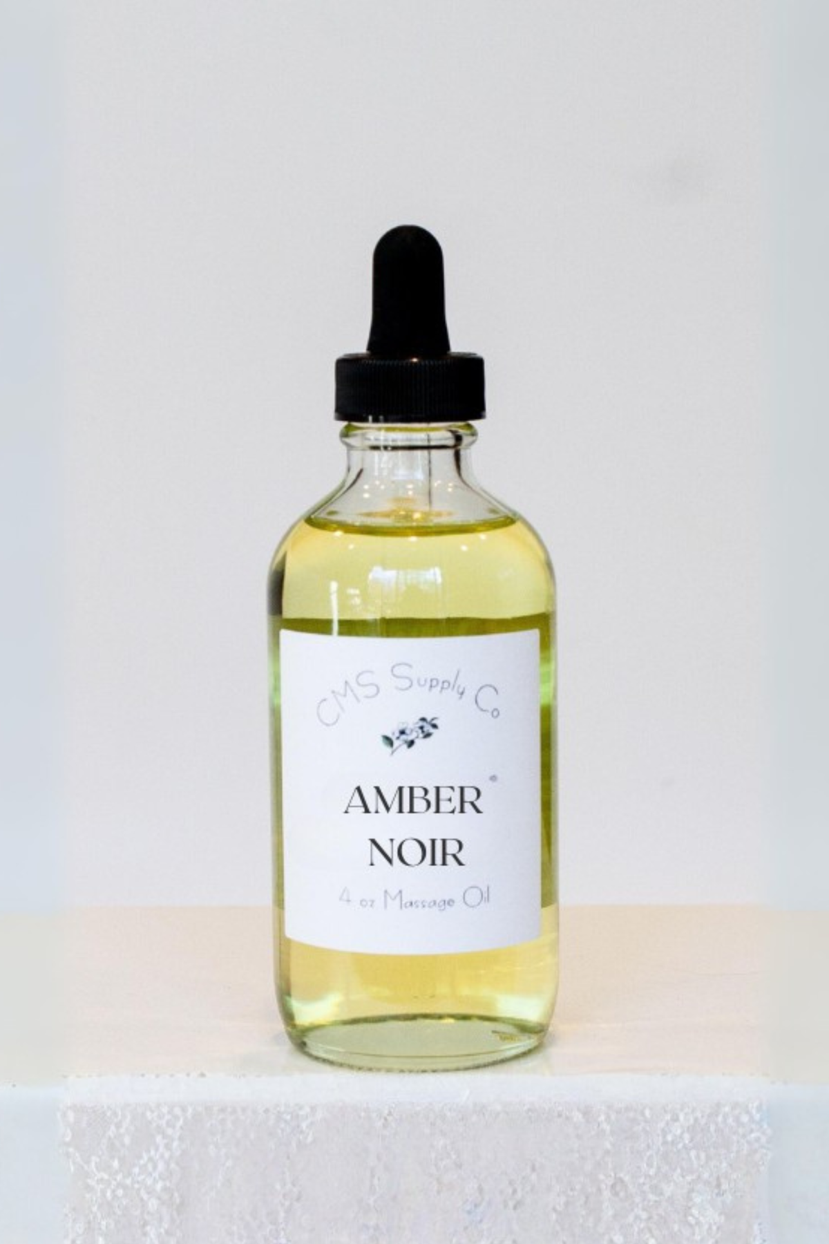 A 4 oz glass dropper bottle of CMS Supply Co Amber Noir massage oil with a white label, standing on a white lace-covered table