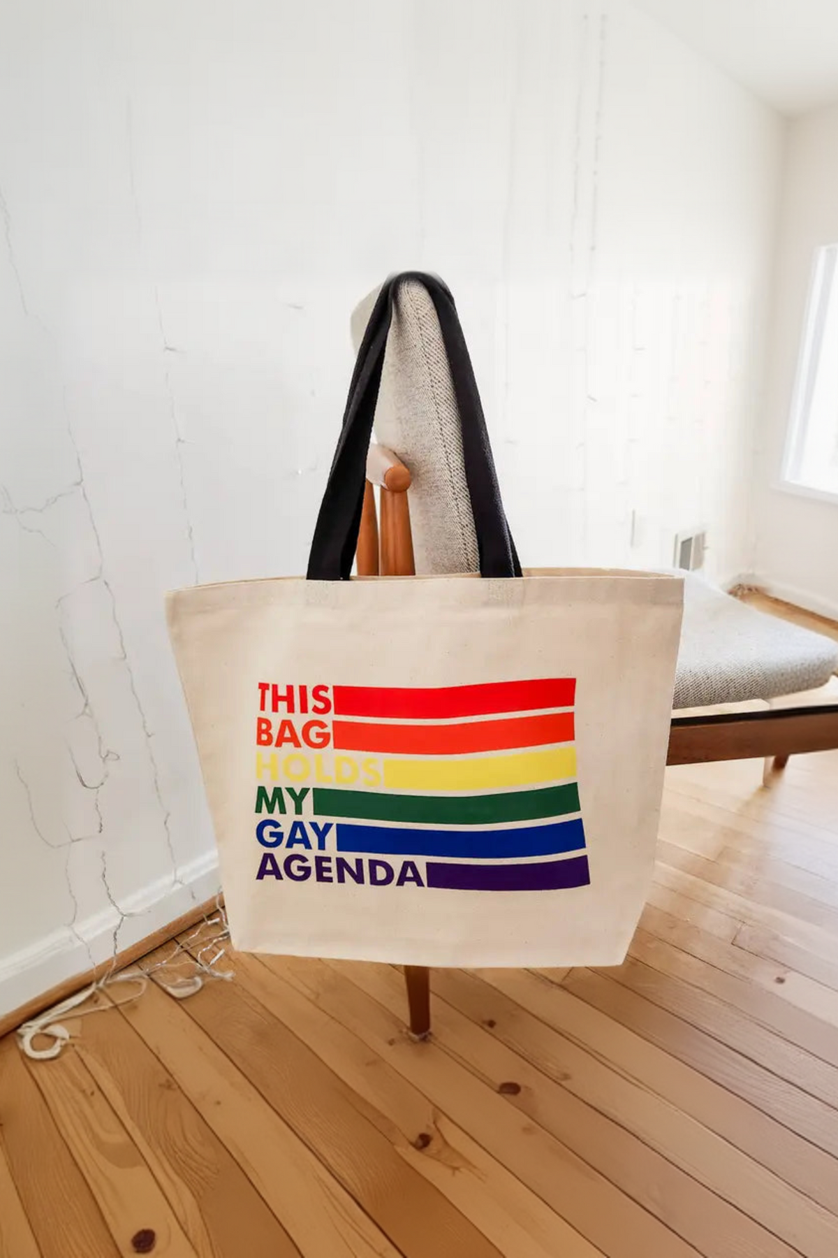 Canvas My Gay Agenda tote hanging by its black handles from a wooden chair in a bright white room, the rainbow striped slogan facing out over a wood floor.