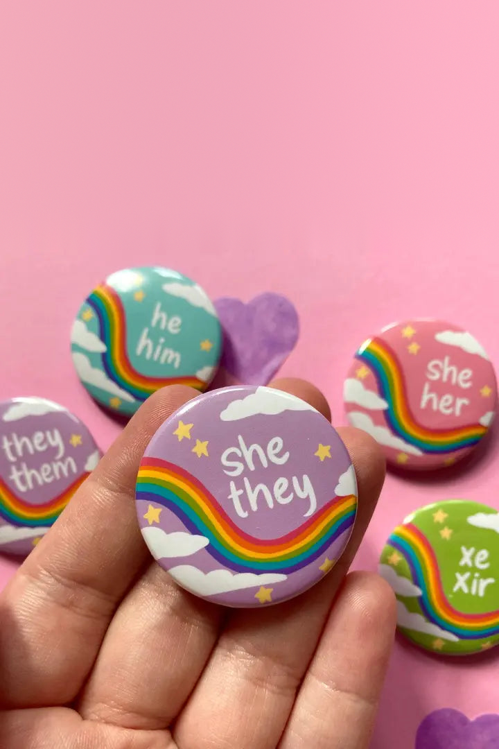 Hand holding a lavender she/they pronoun button with a rainbow and clouds, beside he/him, she/her, they/them and xe/xir pastel buttons on pink
