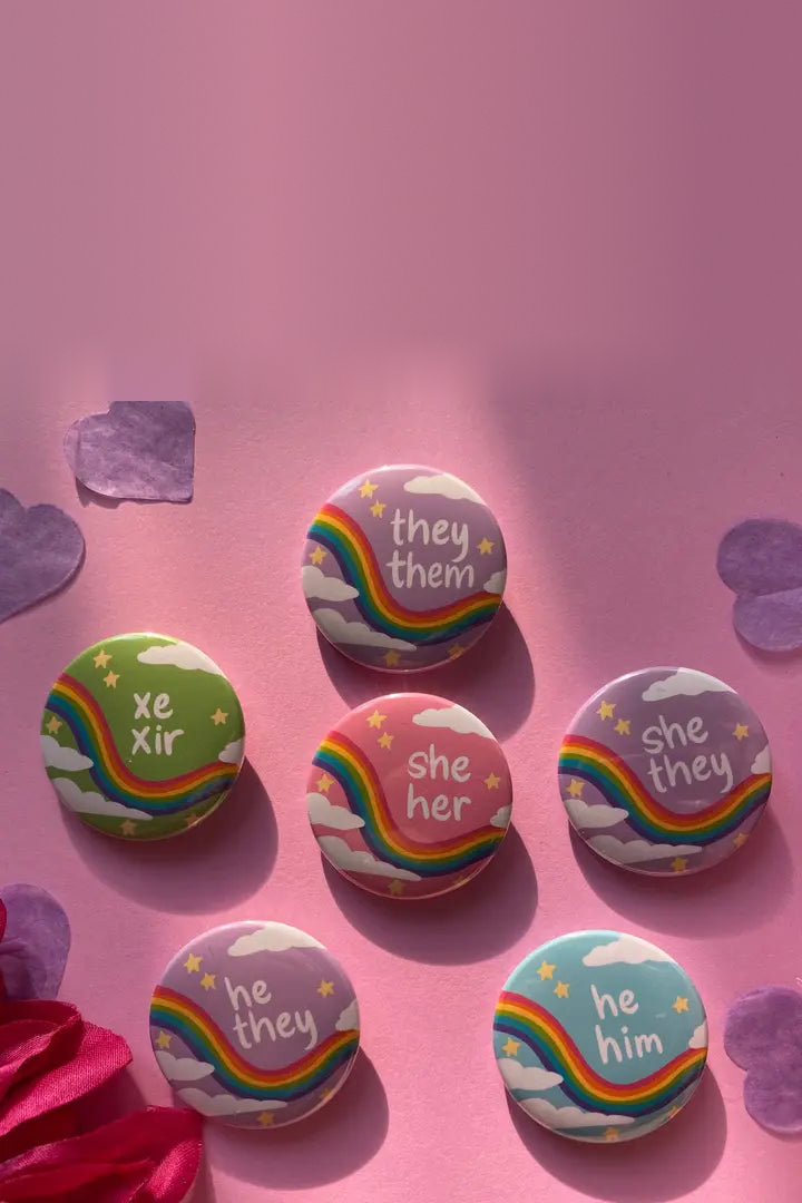 Six pastel rainbow pronoun buttons reading they/them, xe/xir, she/her, she/they, he/they and he/him on pink paper with purple paper hearts